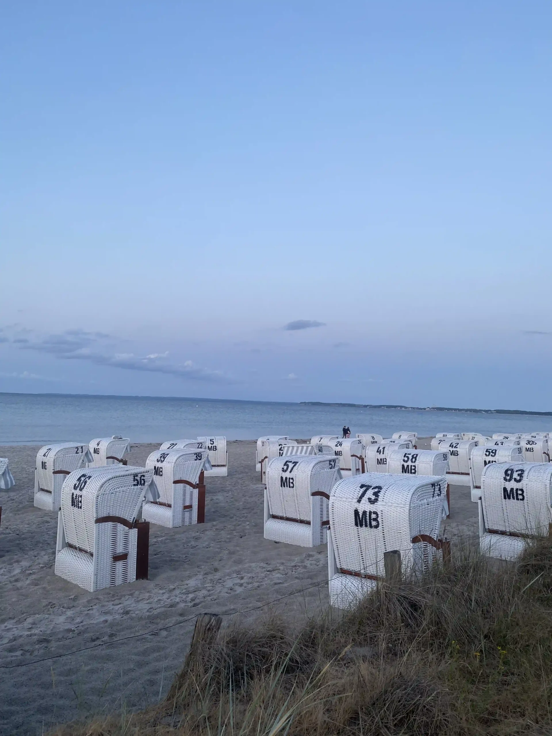 Traditional white hooded beach chairs lined on a sandy Baltic Sea beach at dusk, with calm water, a blue evening sky, and coastal dunes in the foreground.