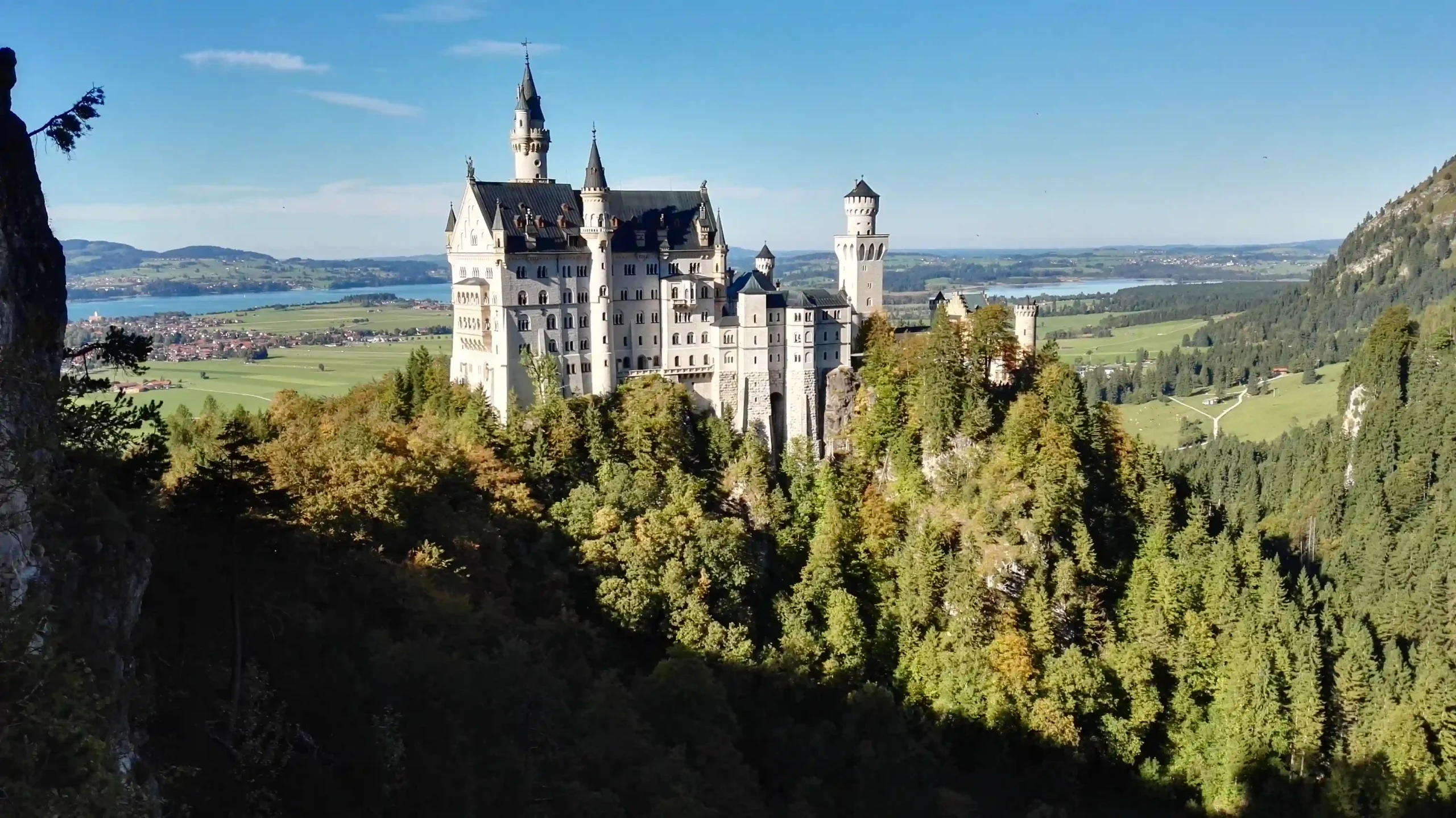 A high-angle scenic view of Neuschwanstein Castle perched on a lush, tree-covered mountain slope with surrounding lakes and valleys.