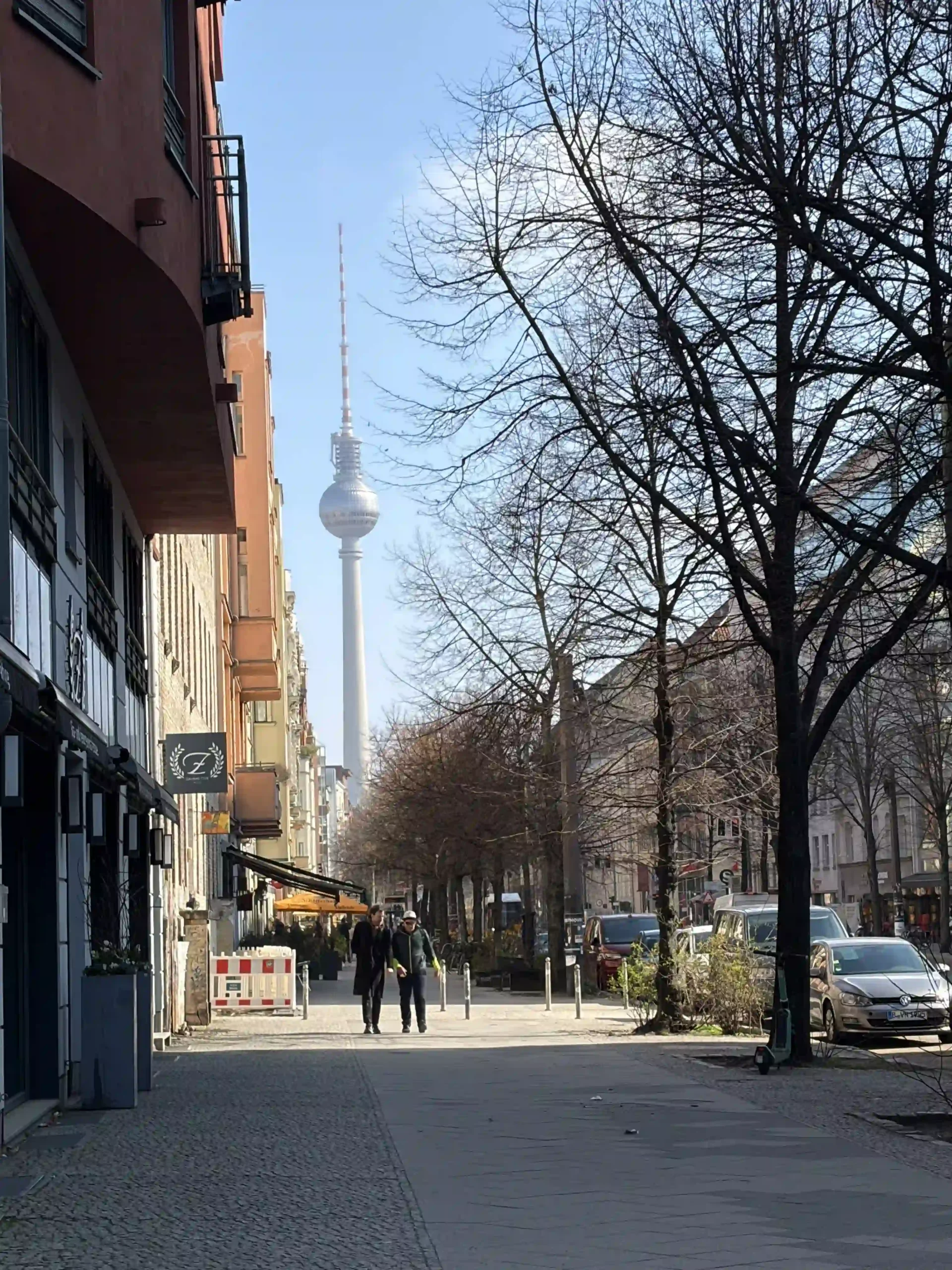 Street view looking down a tree-lined Berlin sidewalk, framed by multi-story city buildings, with the iconic metallic sphere of the Berlin TV Tower towering in the distant background.