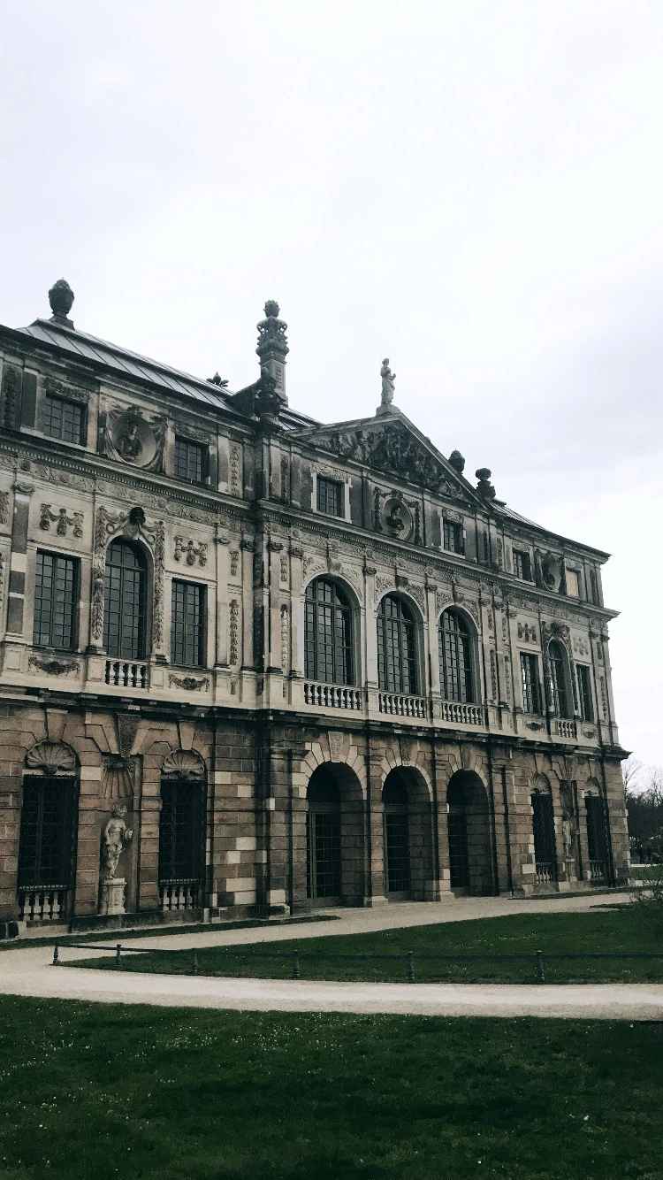 View of the ornate sandstone facade of the Baroque Palais im Großen Garten palace in Dresden, featuring grand arched windows, stone sculptures, and ground-floor arcades next to a green lawn.