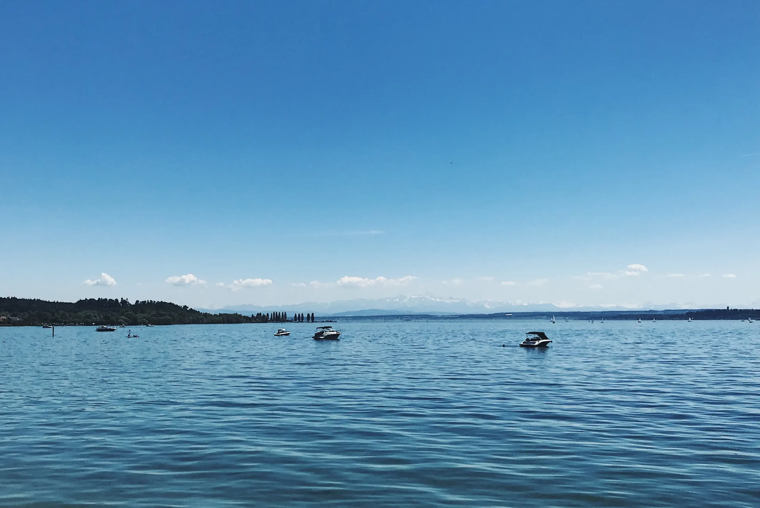 A wide view across the calm blue waters of Lake Constance with several small boats anchored, looking toward faint snow-capped Alps along the distant horizon.