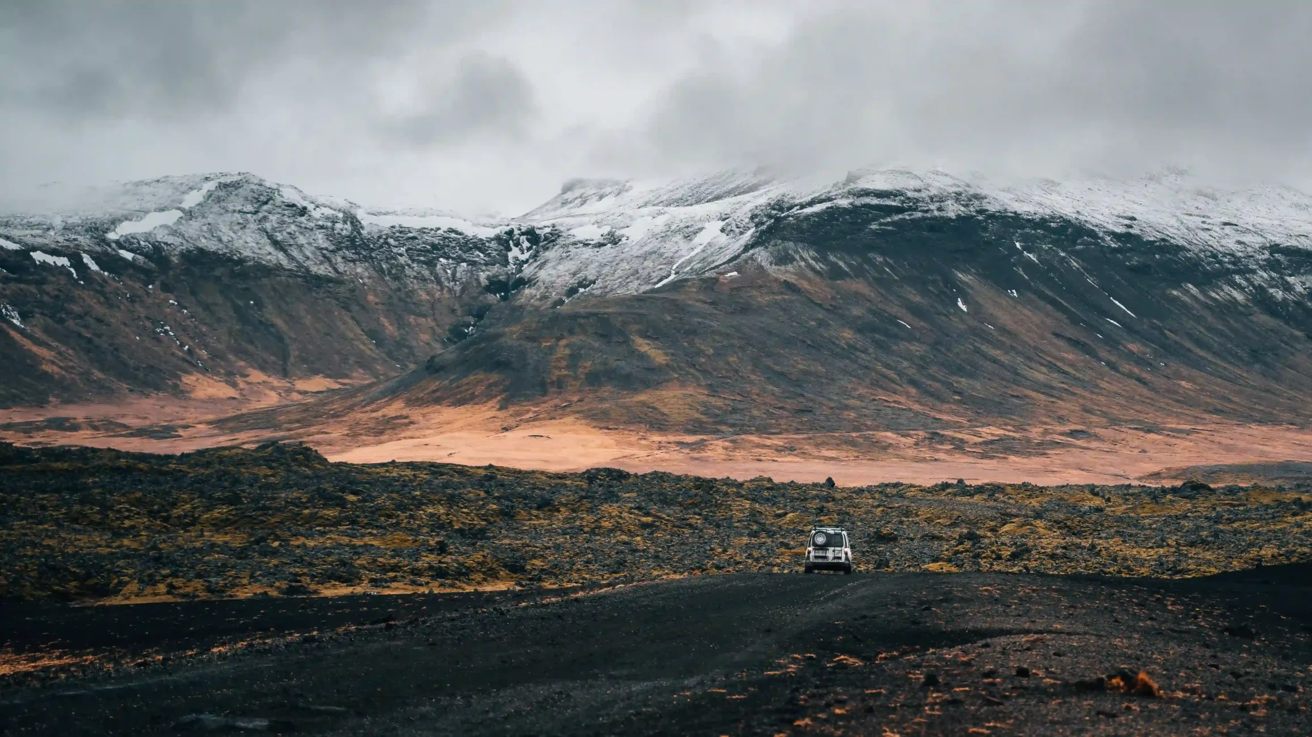 A white camper driving on a dark gravel road through a vast, rugged black lava field toward towering, snow-capped mountains.