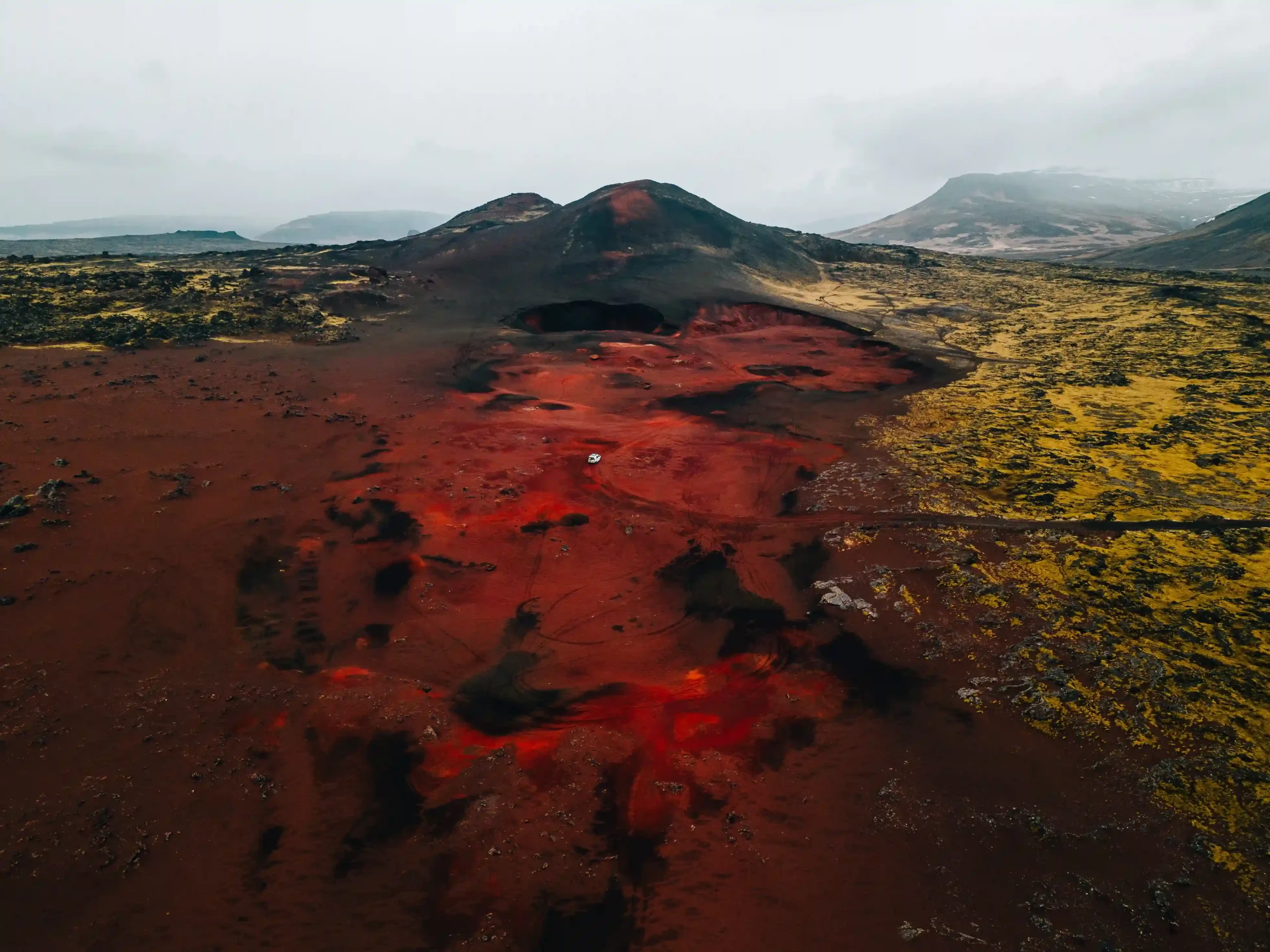 A vibrant red volcanic landscape with a small white camper van parked in the center, surrounded by yellow-green moss and dark mountains under an overcast sky.