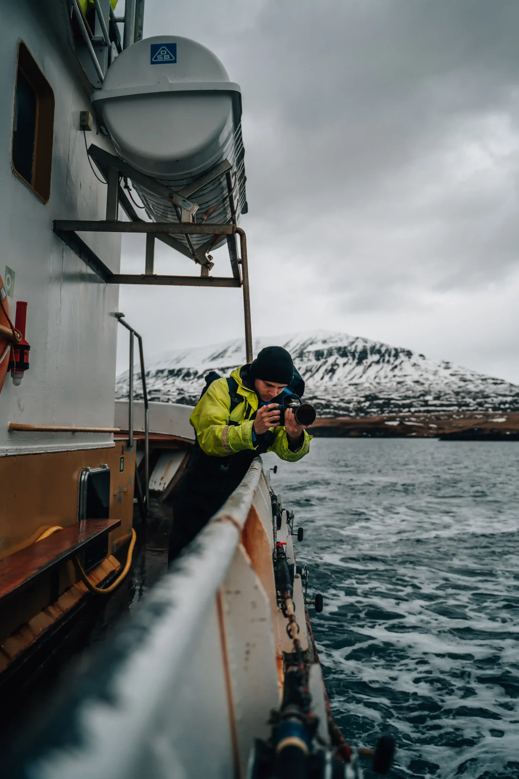 A photographer in a bright yellow jacket leans over the railing of a white boat, pointing a camera toward the dark water.