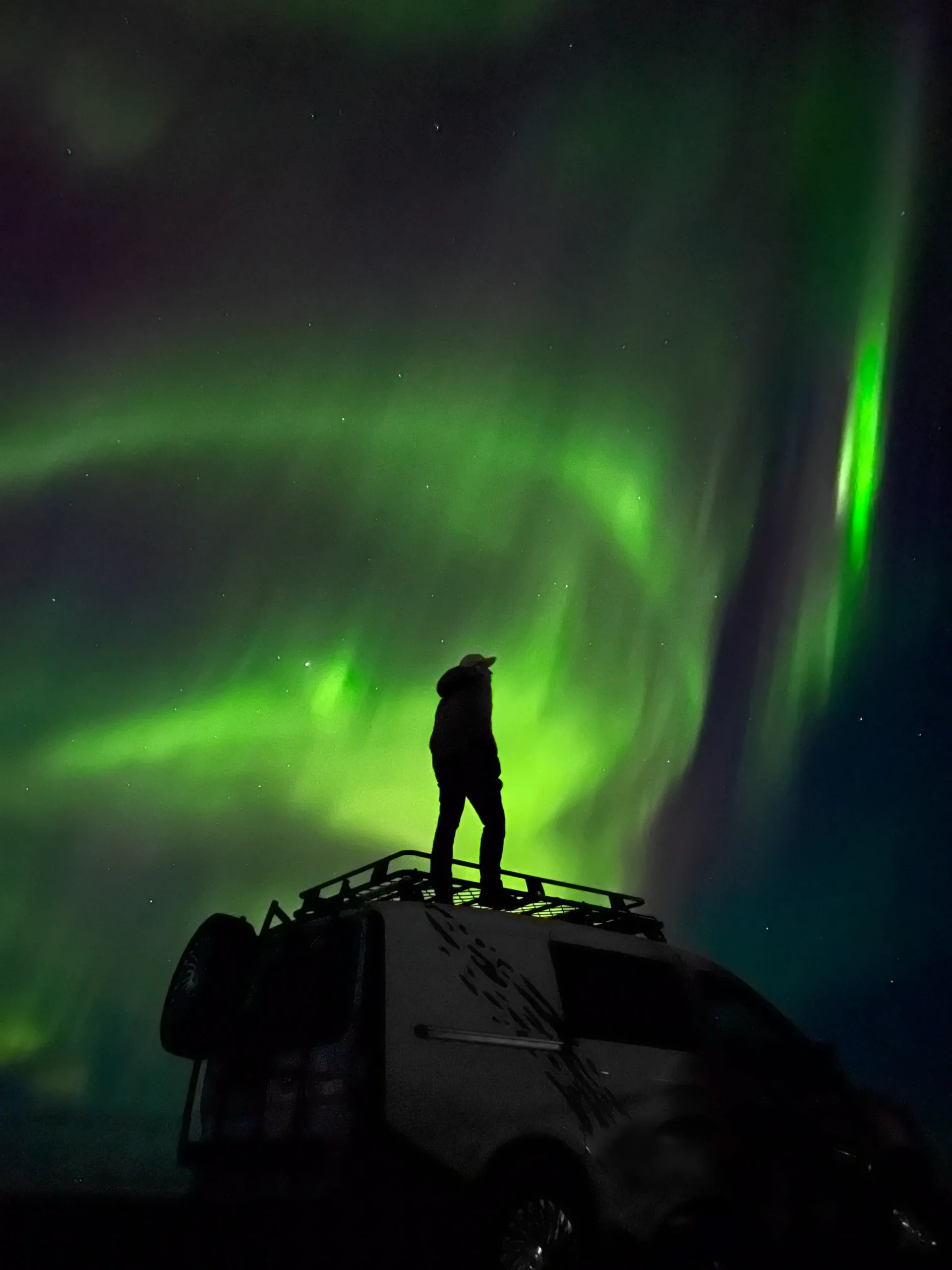 A silhouette of a person standing on the roof rack of a white camper van at night, looking up at vibrant green and purple aurora borealis waves dancing across a starry dark sky.