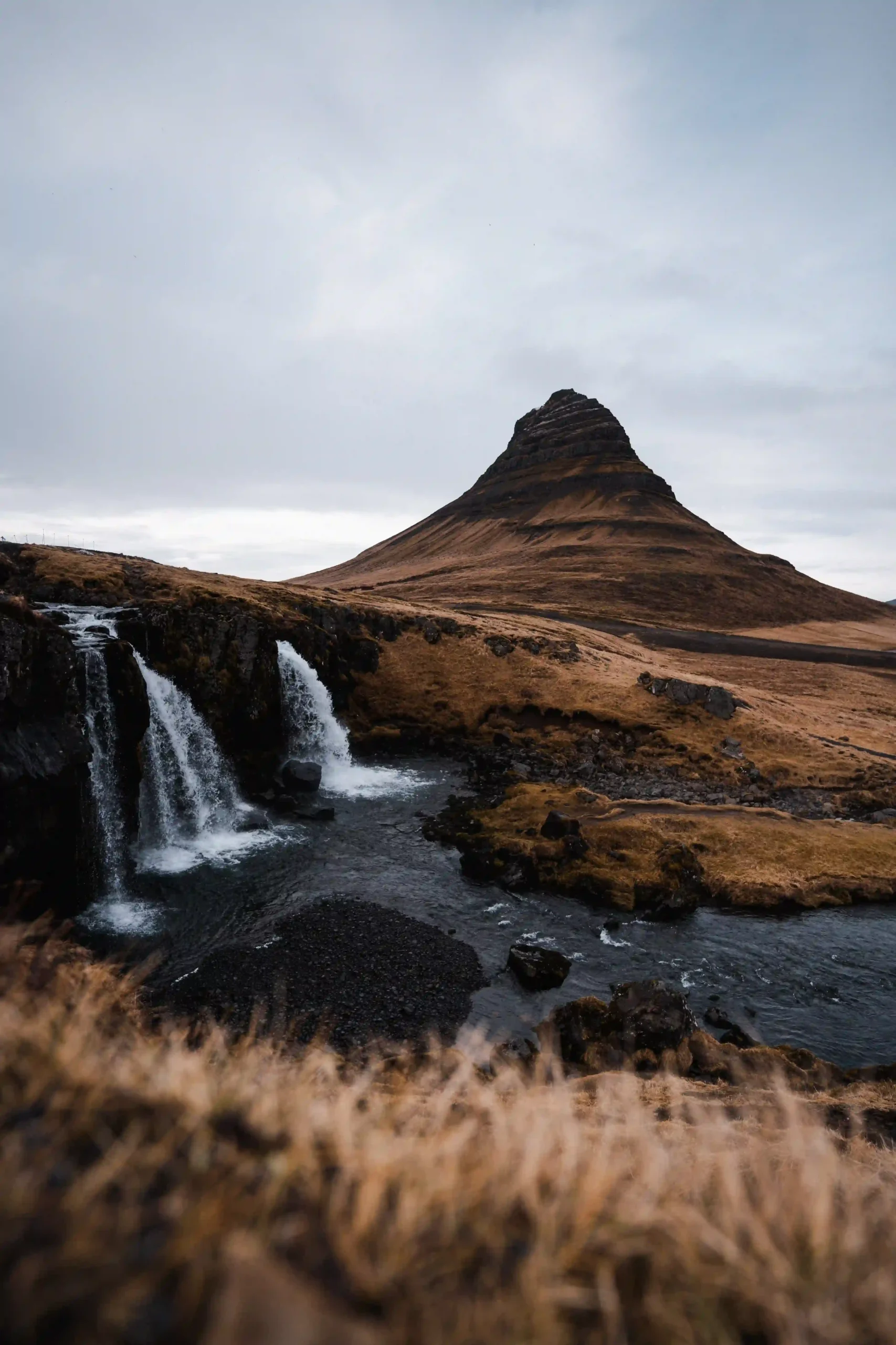 Cone-shaped Kirkjufell mountain and Kirkjufellsfoss waterfall under a cloudy sky.