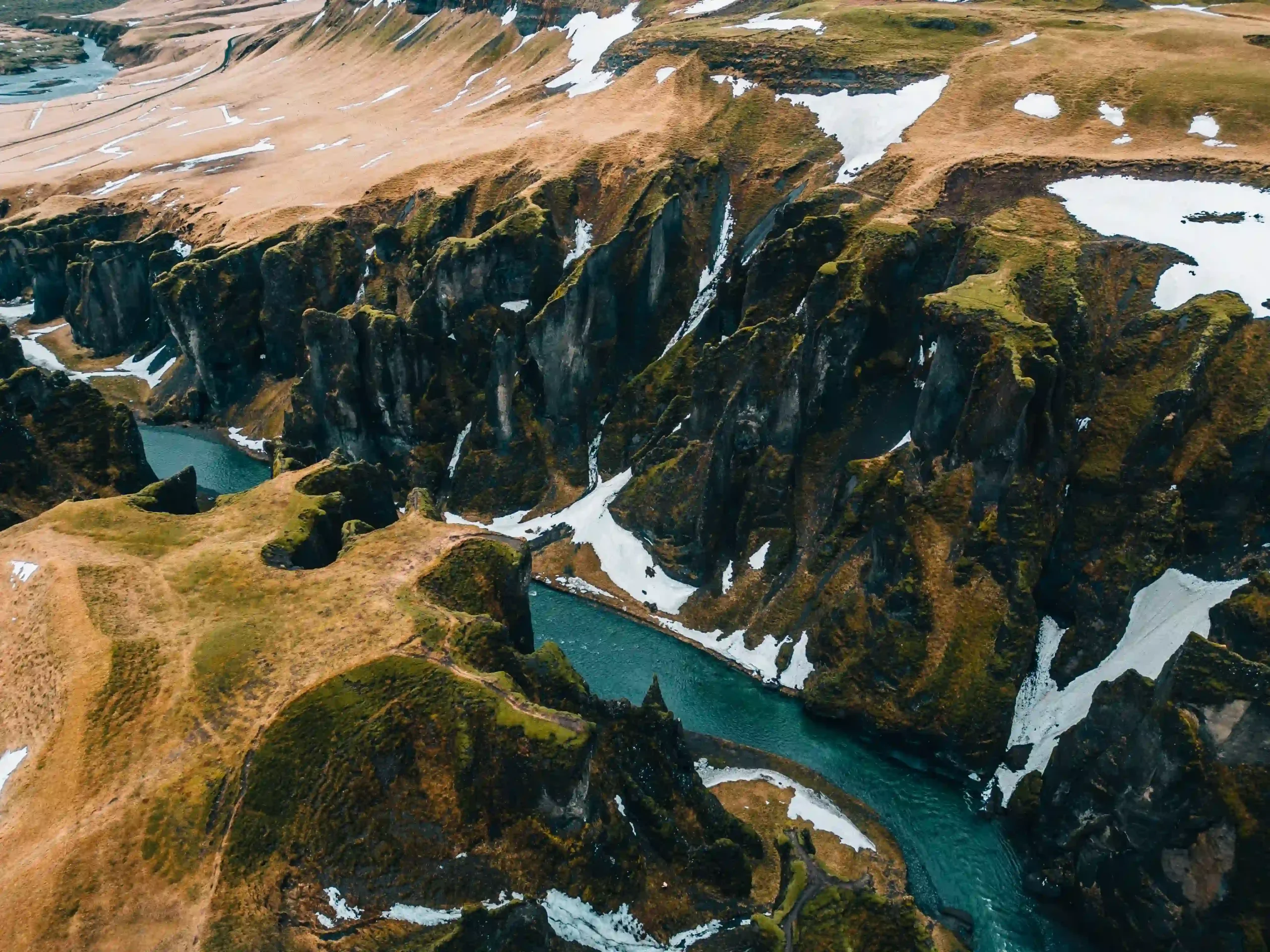 A high-angle drone shot looking down into the deep, winding Fjaðrárgljúfur Canyon, showing a turquoise river flowing between jagged mossy cliffs with patches of snow on the surrounding plains.