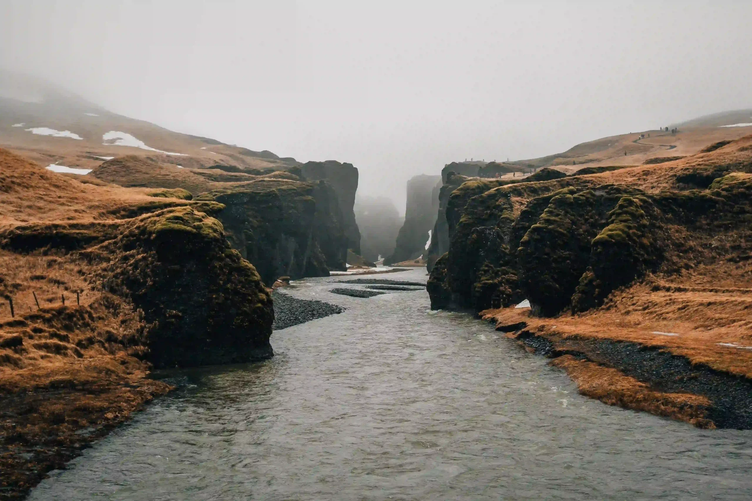 A wide view of a shallow river flowing between towering, moss-covered jagged cliffs of Fjaðrárgljúfur Canyon under a misty, overcast sky.