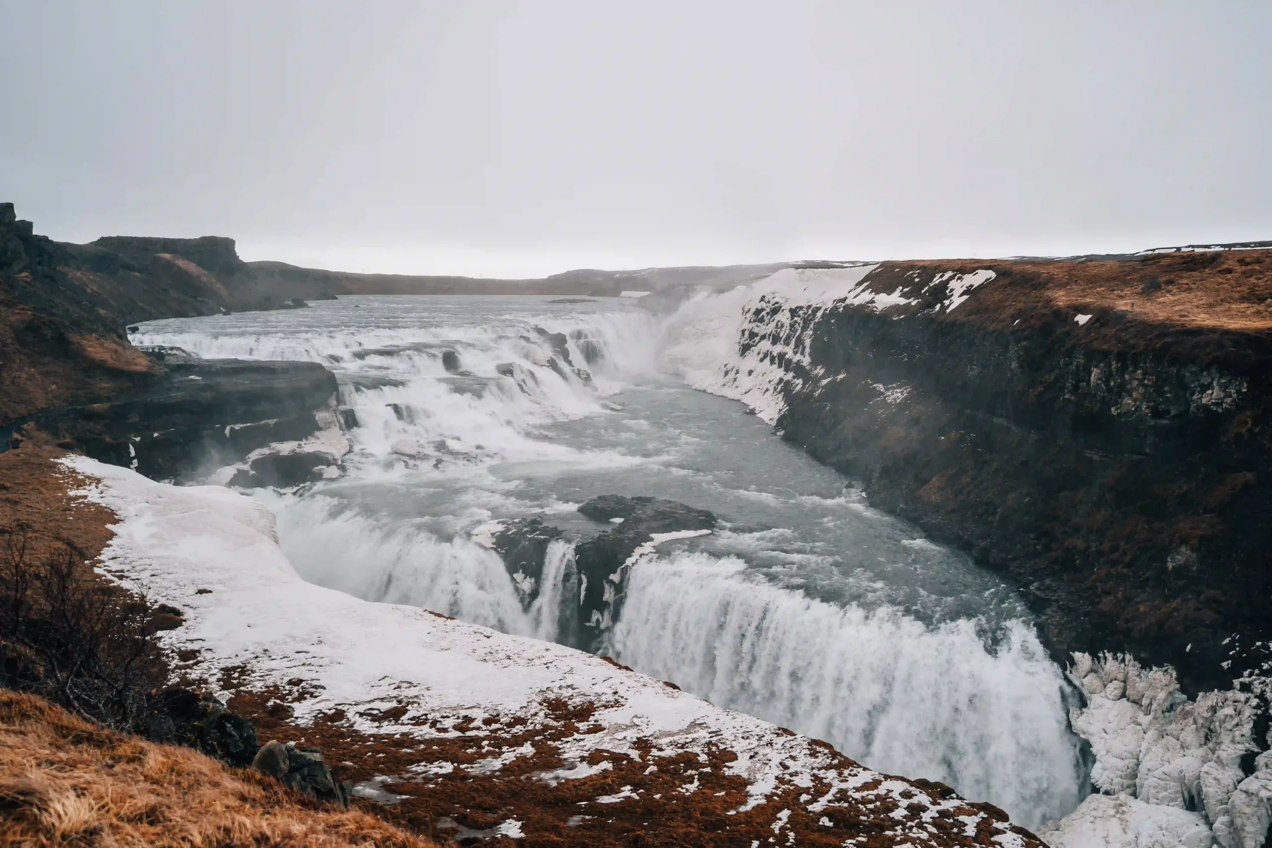 View of Gullfoss waterfall flowing through a rugged canyon with patches of white snow and ice on the dark rocky banks under an overcast sky.