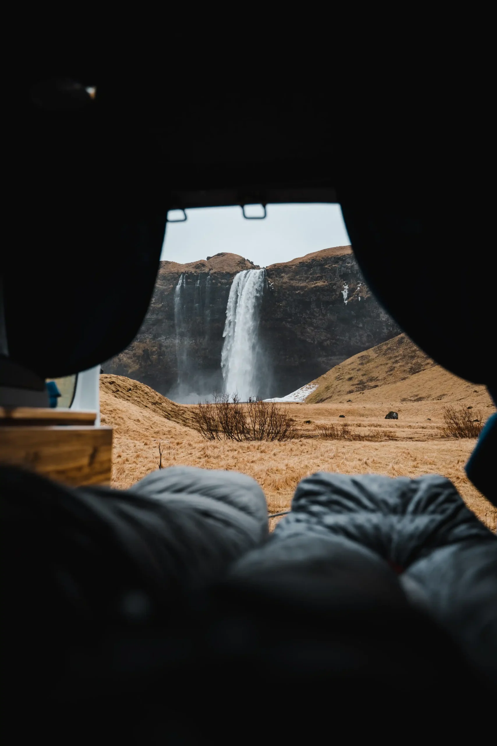 A view from inside a dark camper van looking out through the open back doors at the tall Seljalandsfoss waterfall cascading down a cliff.