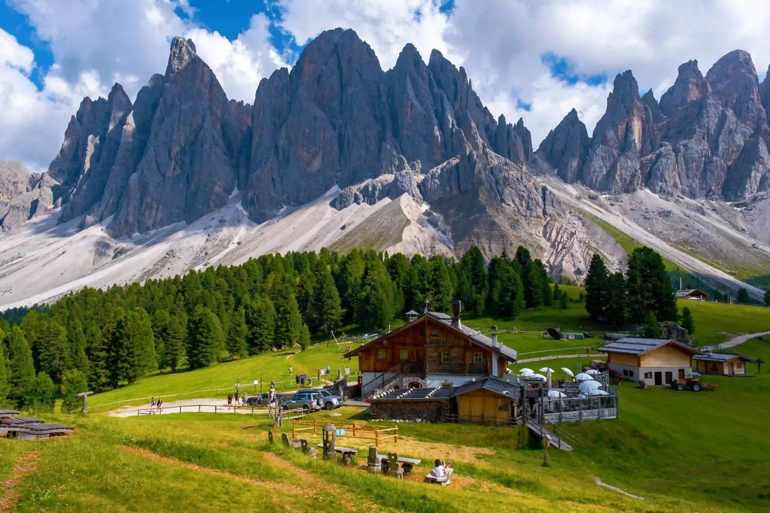 Wide shot of a wooden alpine chalet surrounded by lush green meadows and pine forest, set directly against the towering, jagged rock spires of the Dolomites.