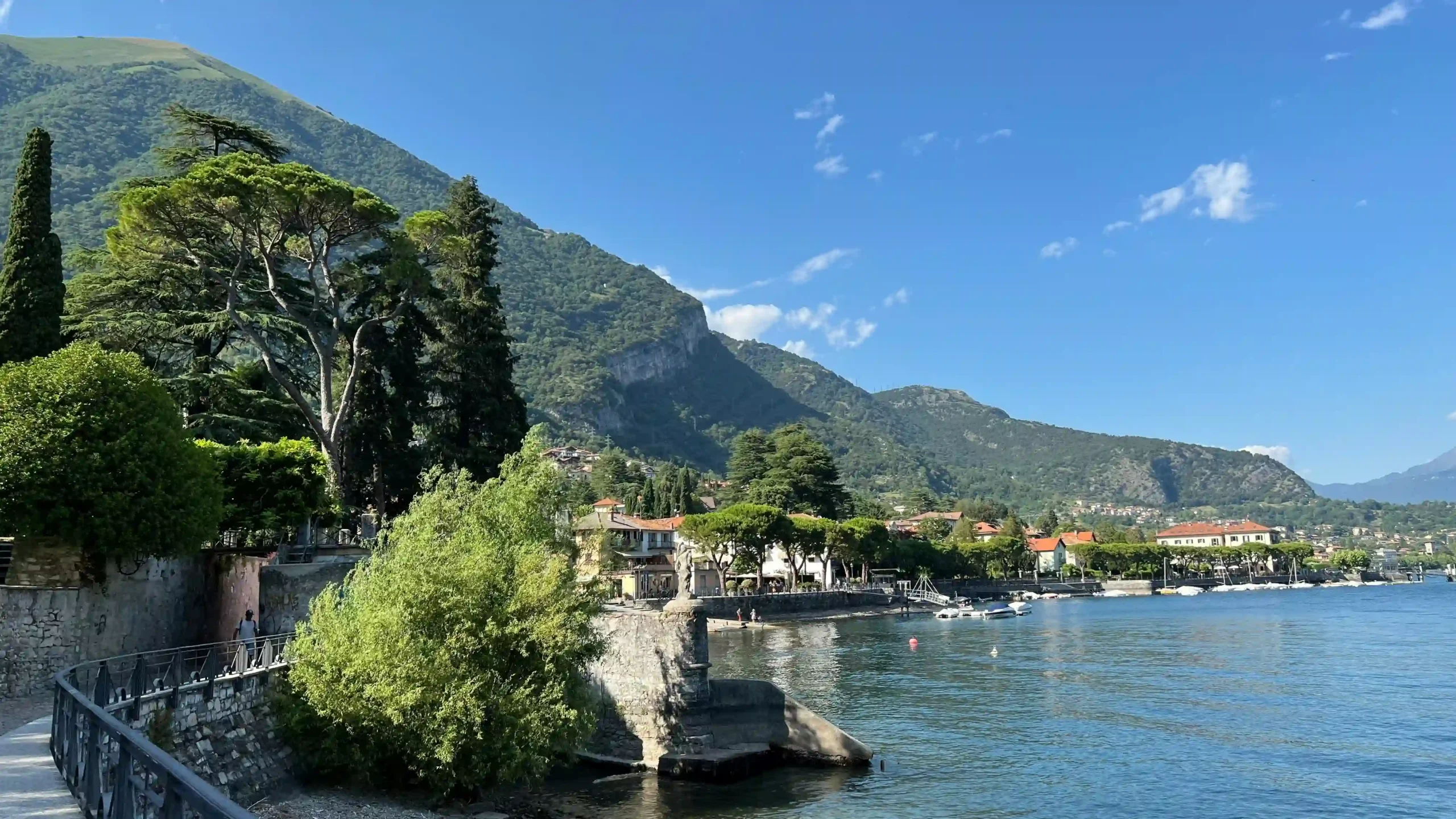 A scenic view of an alpine lake in Italy shows small boats near a coastal town nestled against massive, tree-covered mountain slopes.