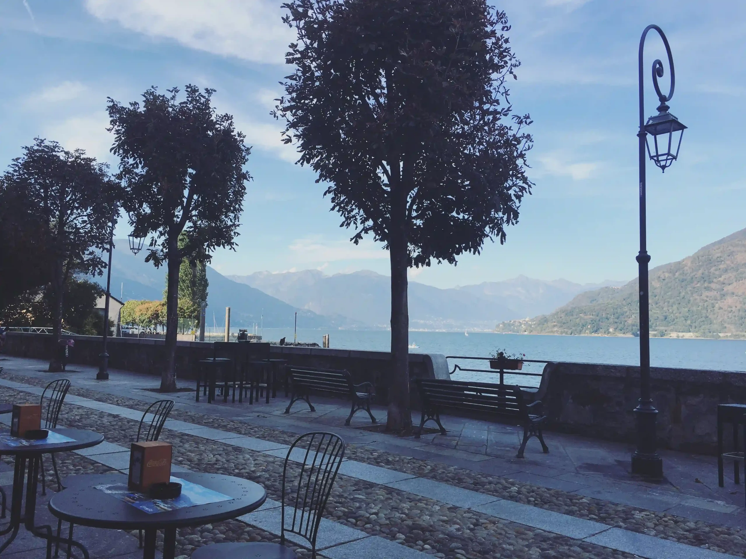 An outdoor cafe terrace with iron tables and chairs on a cobblestone promenade overlooking Lago Maggiore.