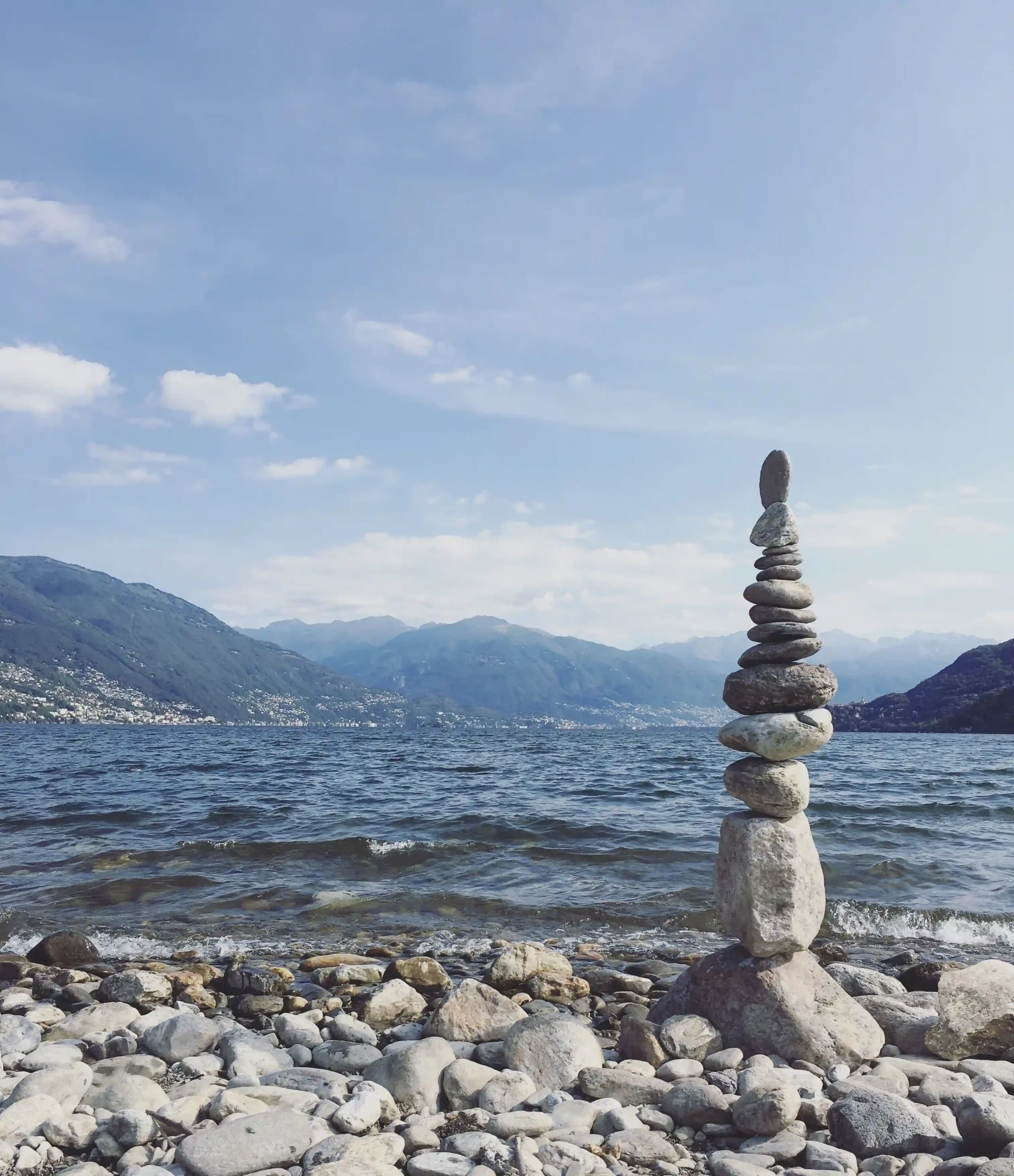 A grey pebble beach, with a rock stack, overlooking the wide, blue expanse of Lago Maggiore.
