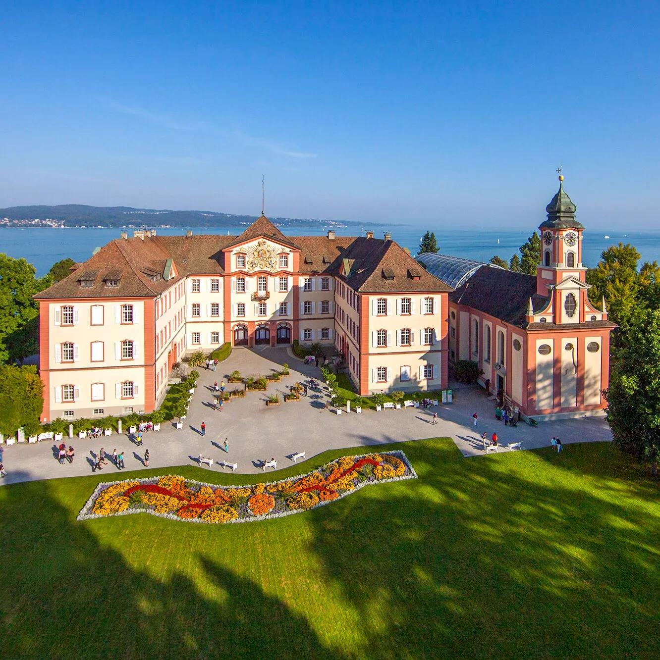 An aerial view of the pink and cream Baroque castle and church on Mainau Island, surrounded by green lawns and flower beds with Lake Constance in the background.