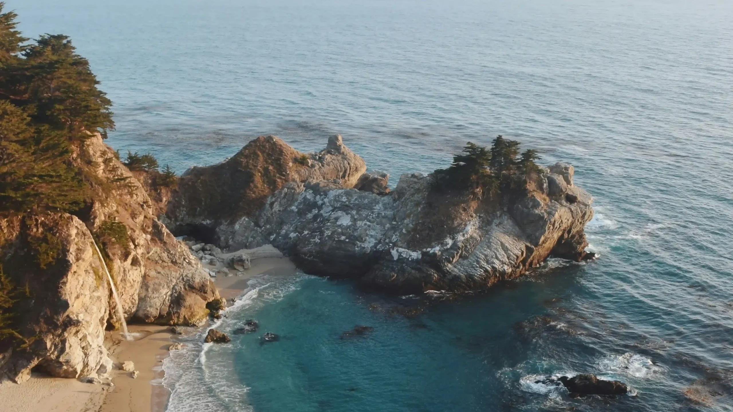 View of McWay Falls, a waterfall dropping onto a sandy beach in a turquoise cove, surrounded by jagged cliffs and the vast Pacific Ocean under a soft morning sky in Big Sur.