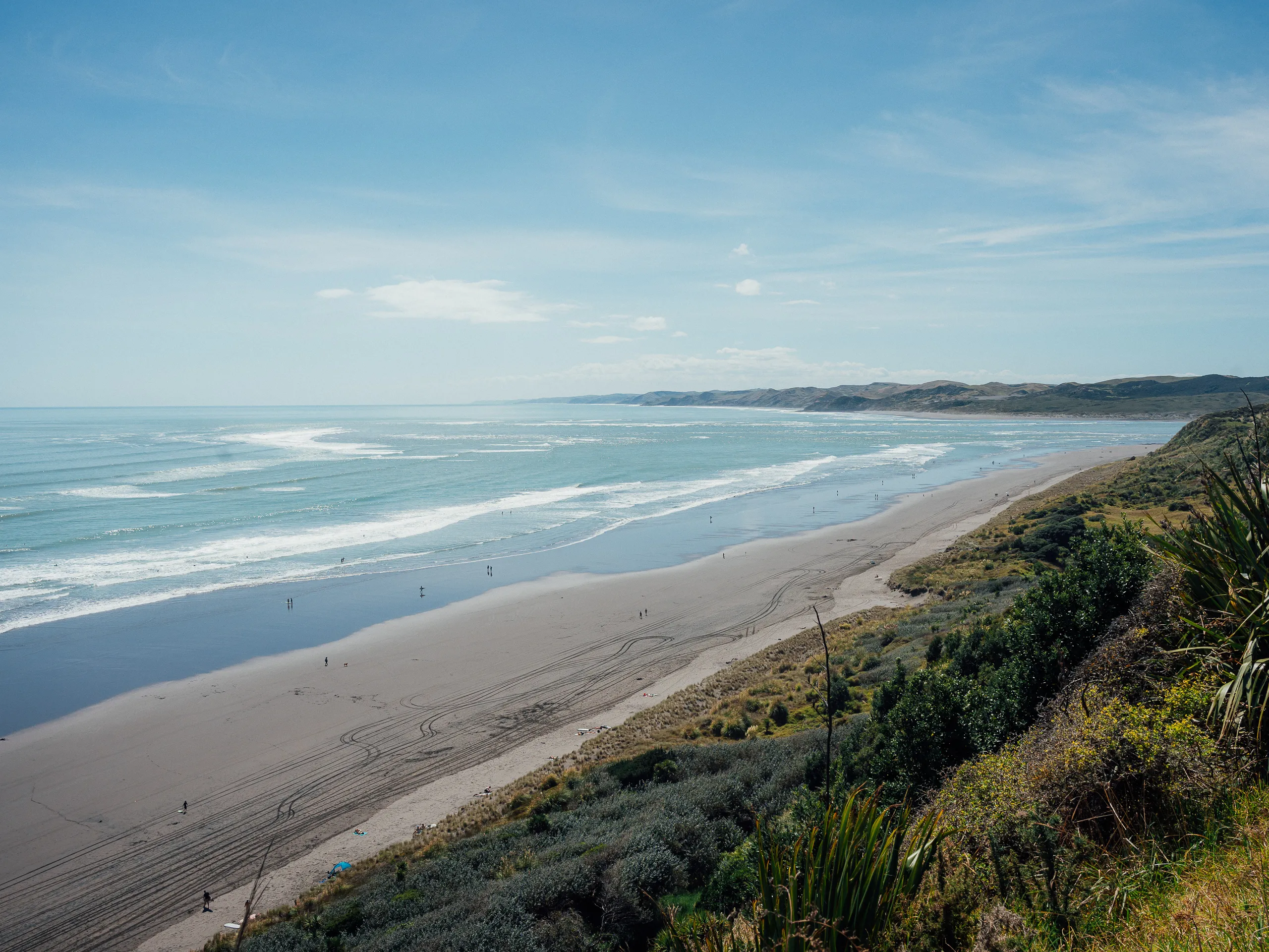 A long, dark-sand beach meeting a turquoise ocean, with green coastal cliffs and tracks in the sand.