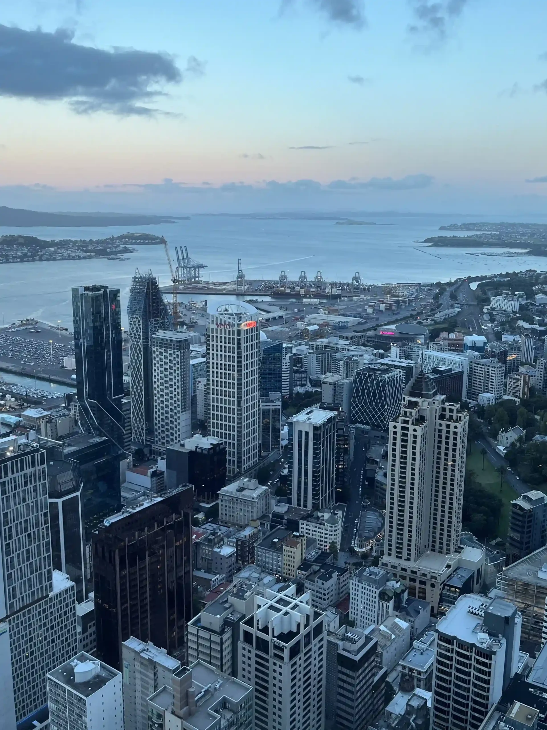 A high-angle aerial view of Auckland’s skyline featuring modern skyscrapers, the city harbor with shipping cranes, and distant islands under a soft dusk sky.
