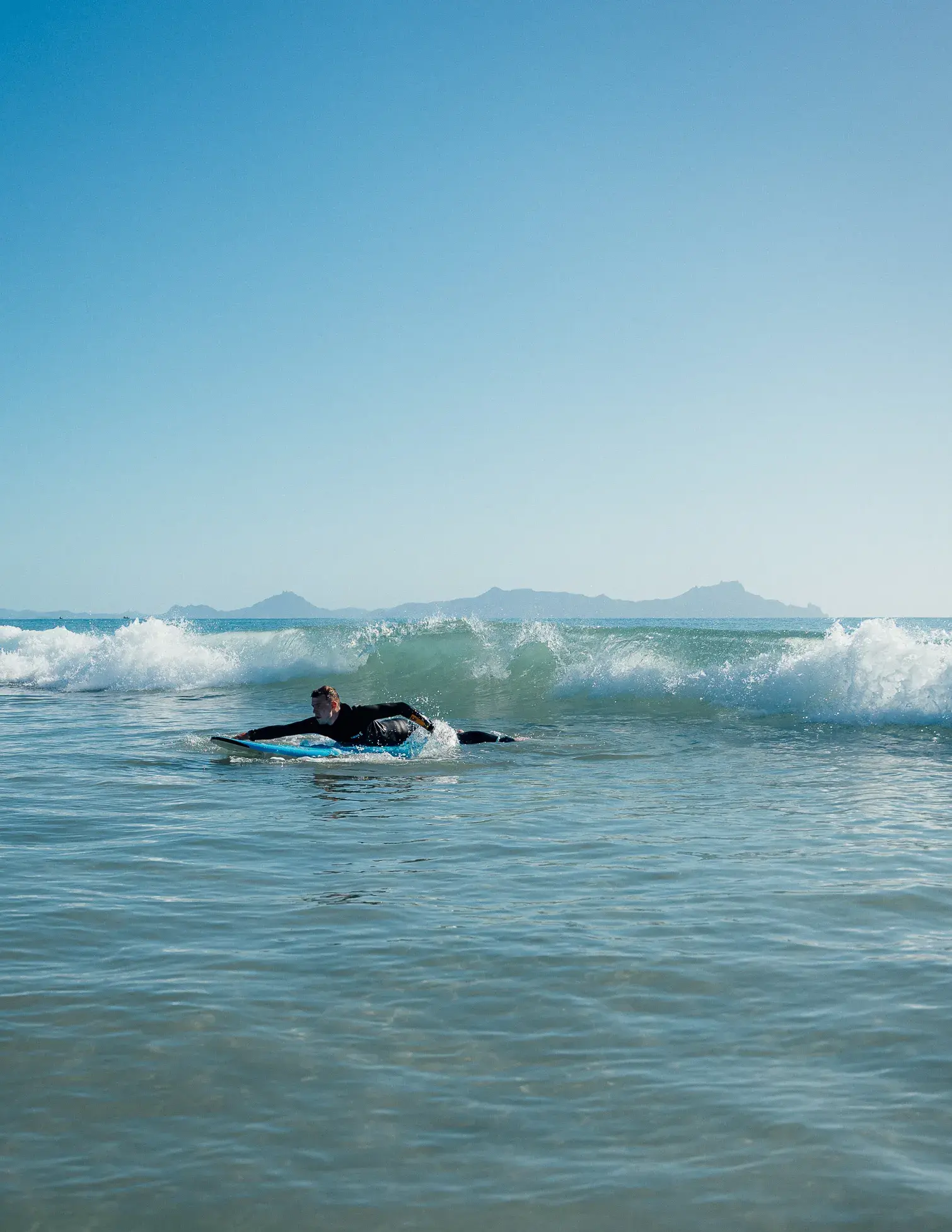 A person in a black wetsuit on a blue surfboard paddling through a clear ocean wave with mountains visible on the horizon under a bright blue sky.