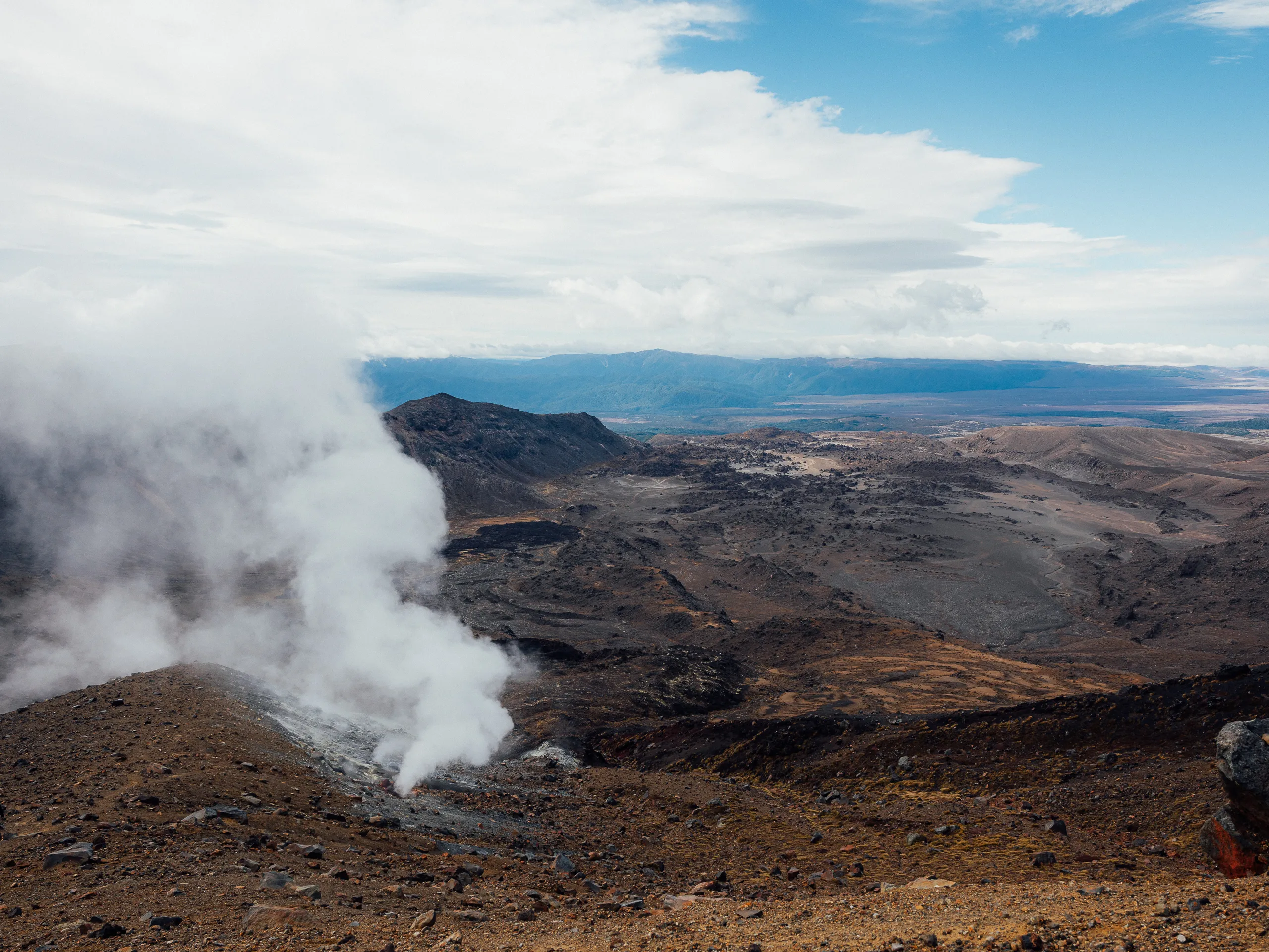 A rugged, volcanic landscape of dark rock and brown earth with white steam rising from a geothermal vent.