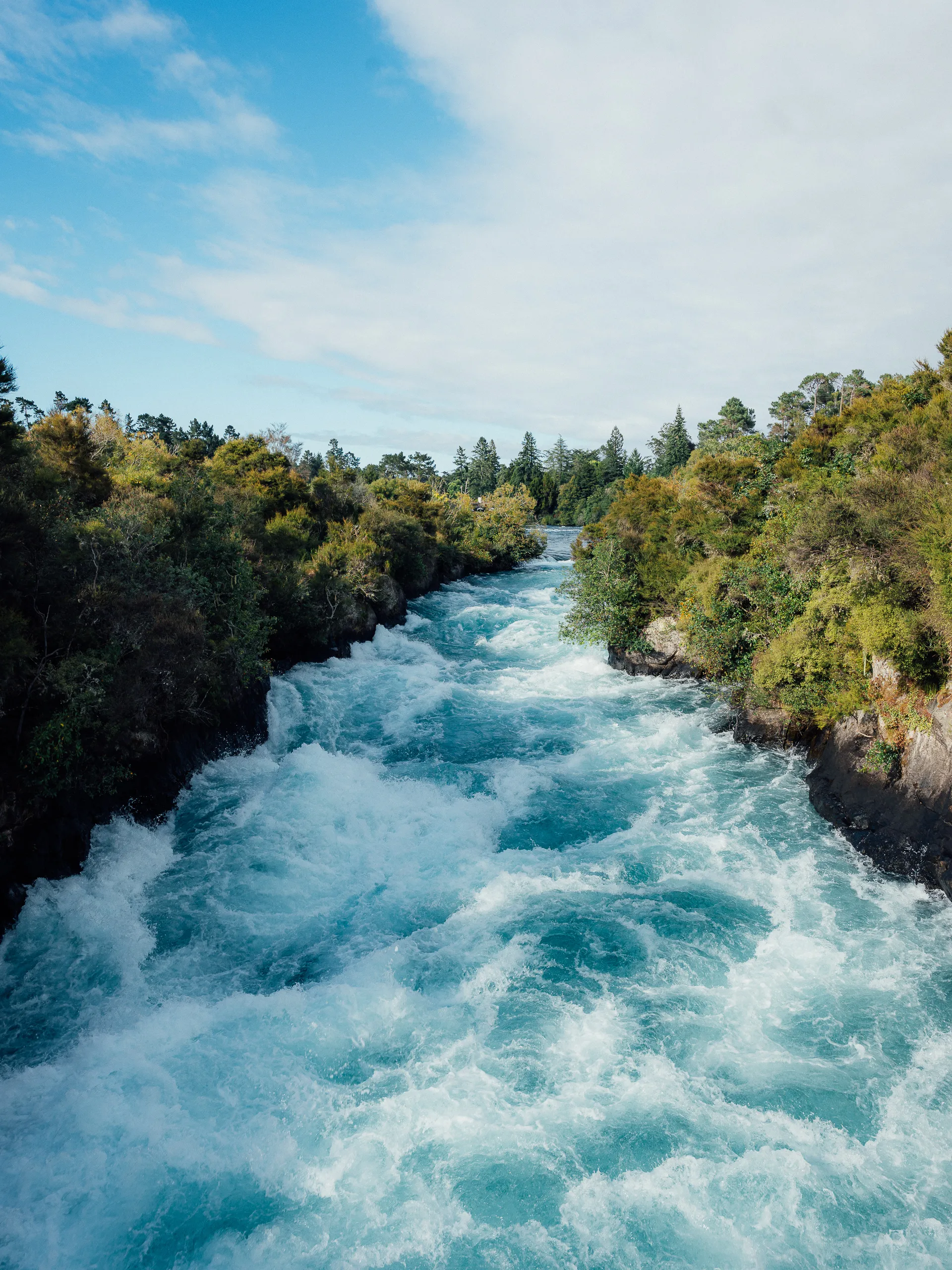 A powerful river of churning, bright turquoise white-water rapids rushing through a narrow rocky channel lined with green trees.