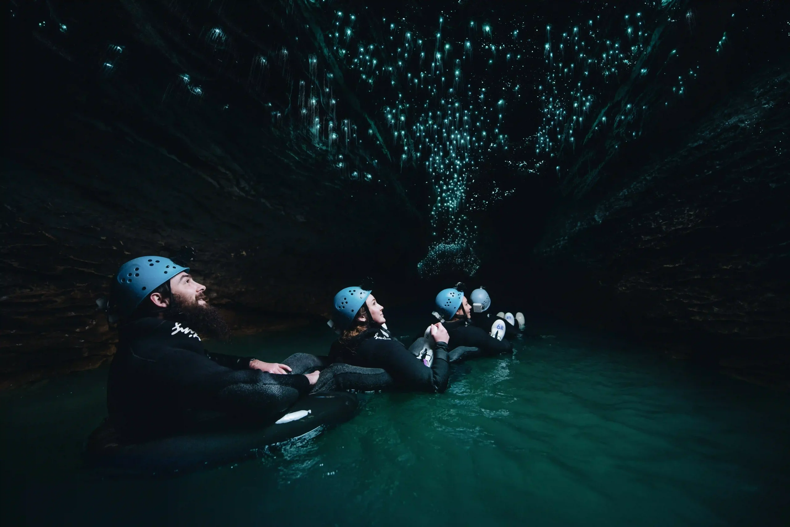 People in helmets and wetsuits floating on an underground river, looking up at a cave ceiling covered in tiny, glowing blue lights.