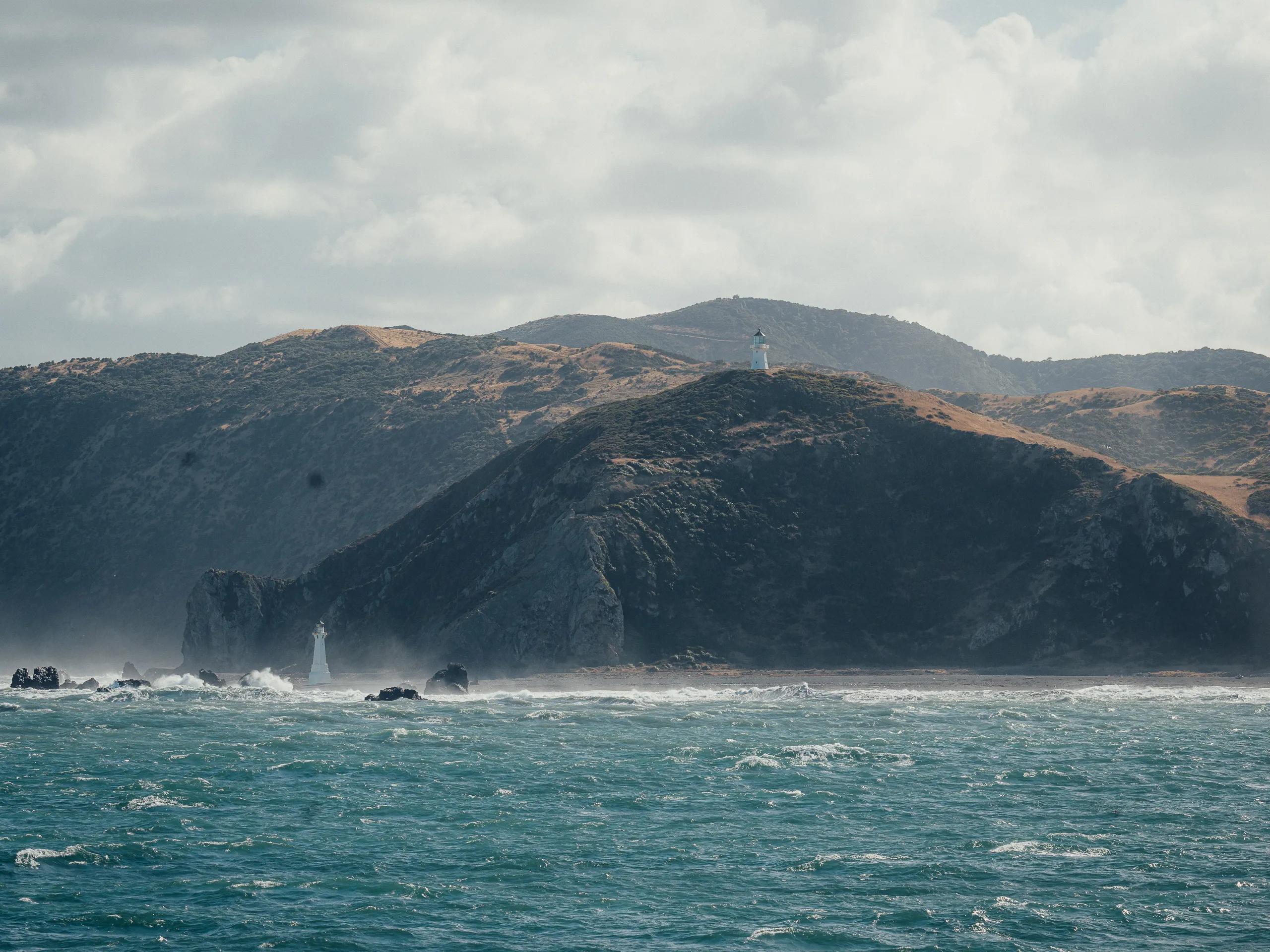 A view across choppy blue water toward brown coastal hills featuring two white lighthouses—one on a high ridge and one at the shoreline.