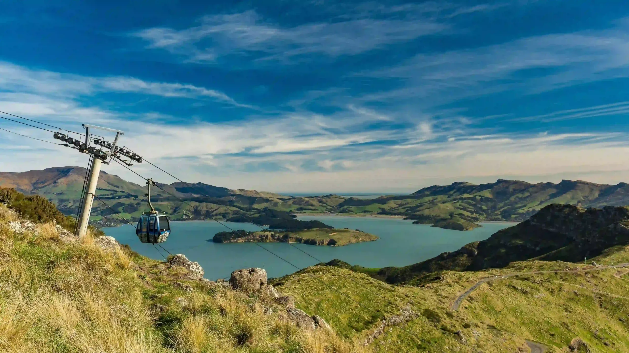 A blue gondola cabin suspended on a cable over green hills, with a view of a turquoise harbor.