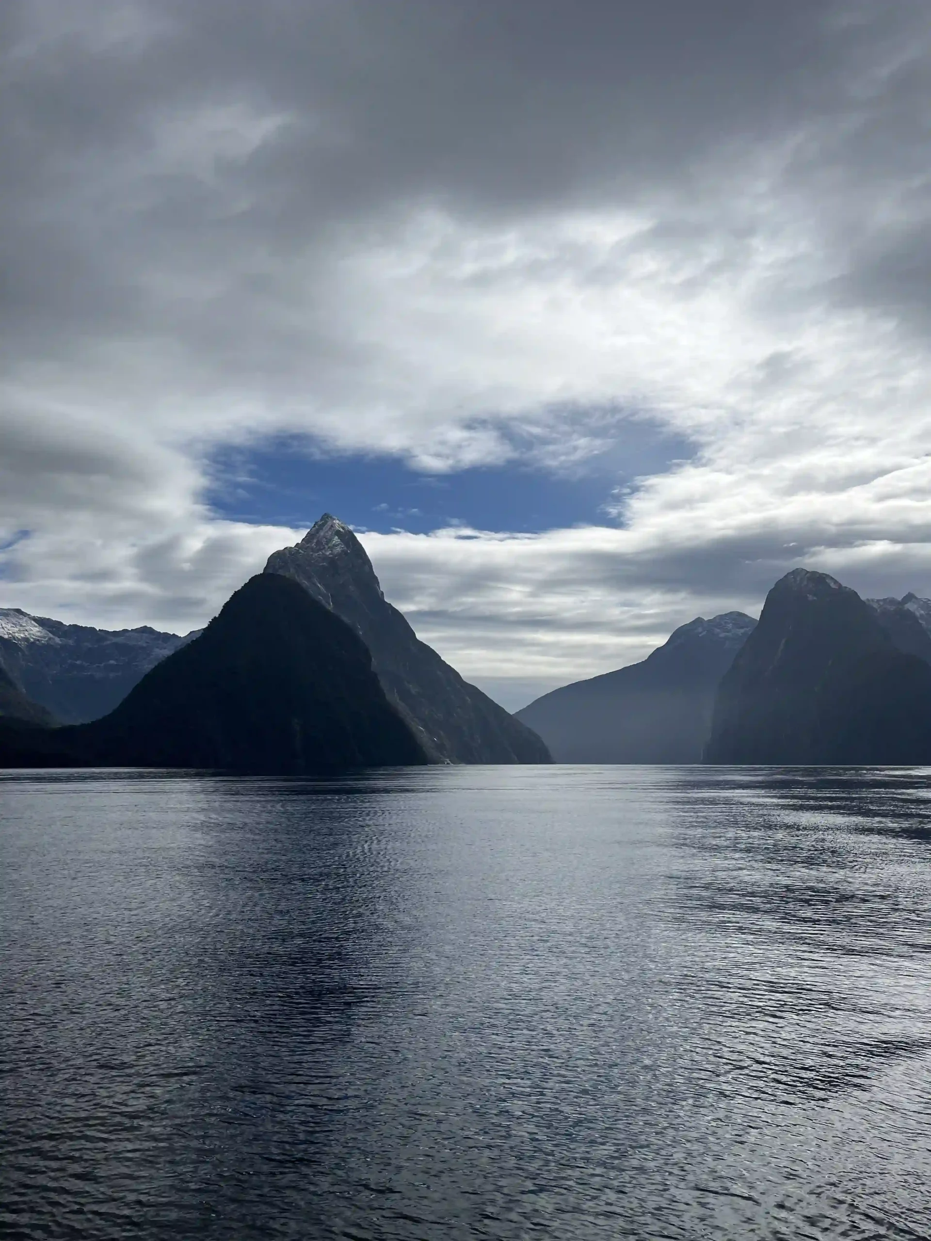 Mitre Peak rising from the still waters of a fjord under a cloudy sky.