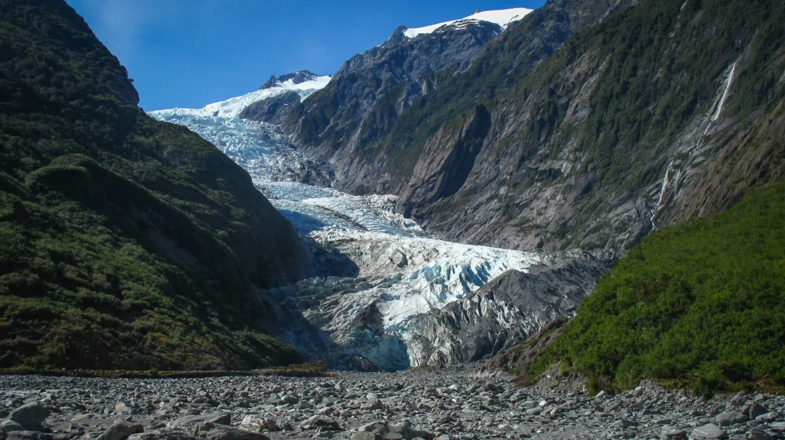 A view looking up a rocky river valley toward a massive white and blue glacier flowing down between steep, green mountainsides.