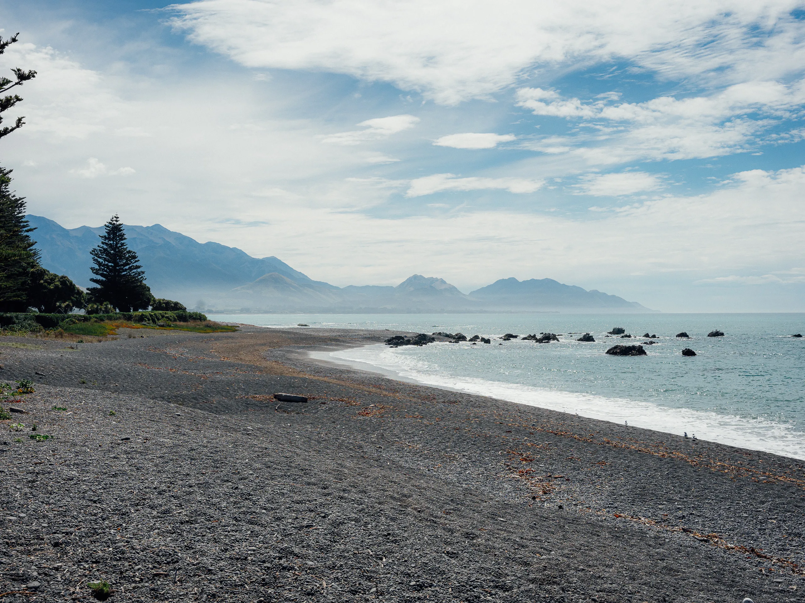 A wide view of a dark grey pebble beach curving toward distant, mist-covered blue mountains under a bright, cloudy sky.