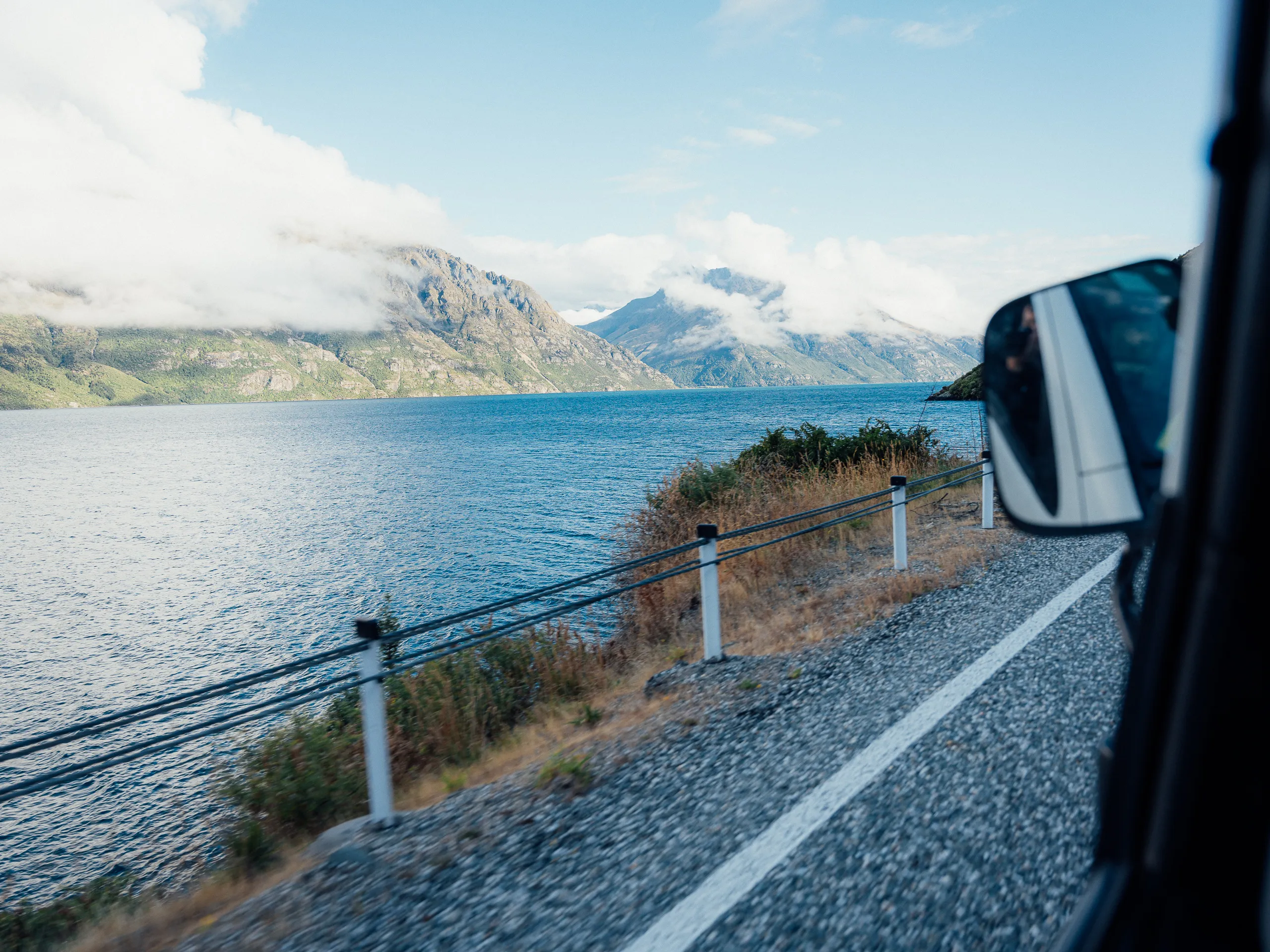 View from a vehicle window looking out over a deep blue lake and cloud-capped mountains.
