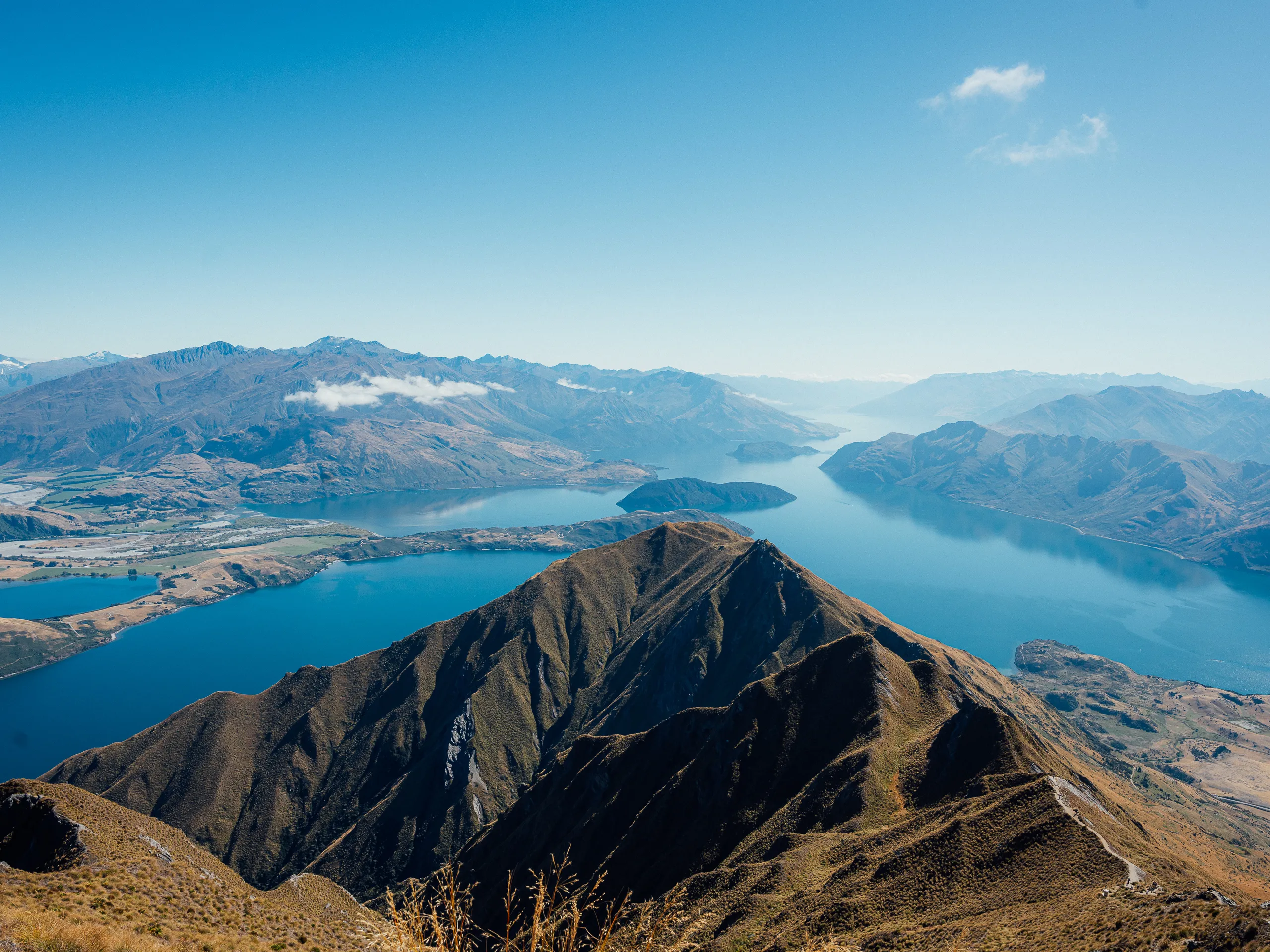 A high-altitude view looking down a sharp, brown alpine ridge toward the expansive blue waters of Lake Wānaka and surrounding mountain peaks.