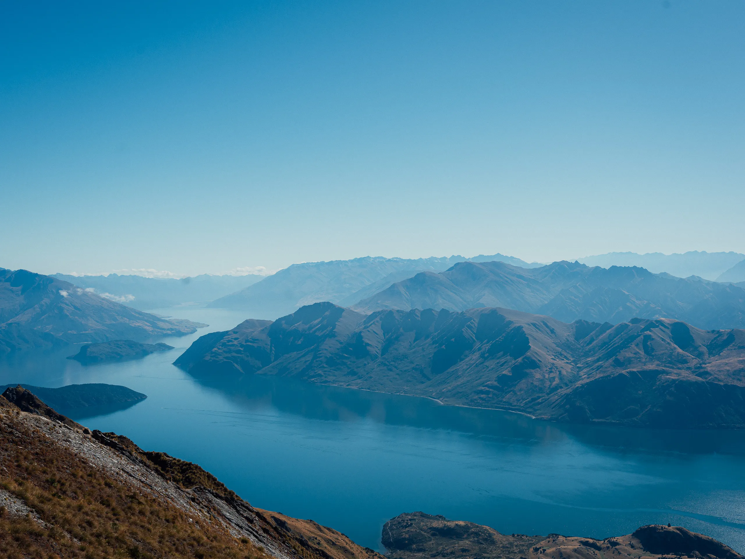 A high-altitude landscape view from a steep mountain ridge looking down at a deep blue lake winding through rugged, sun-drenched mountain ranges under a clear sky.