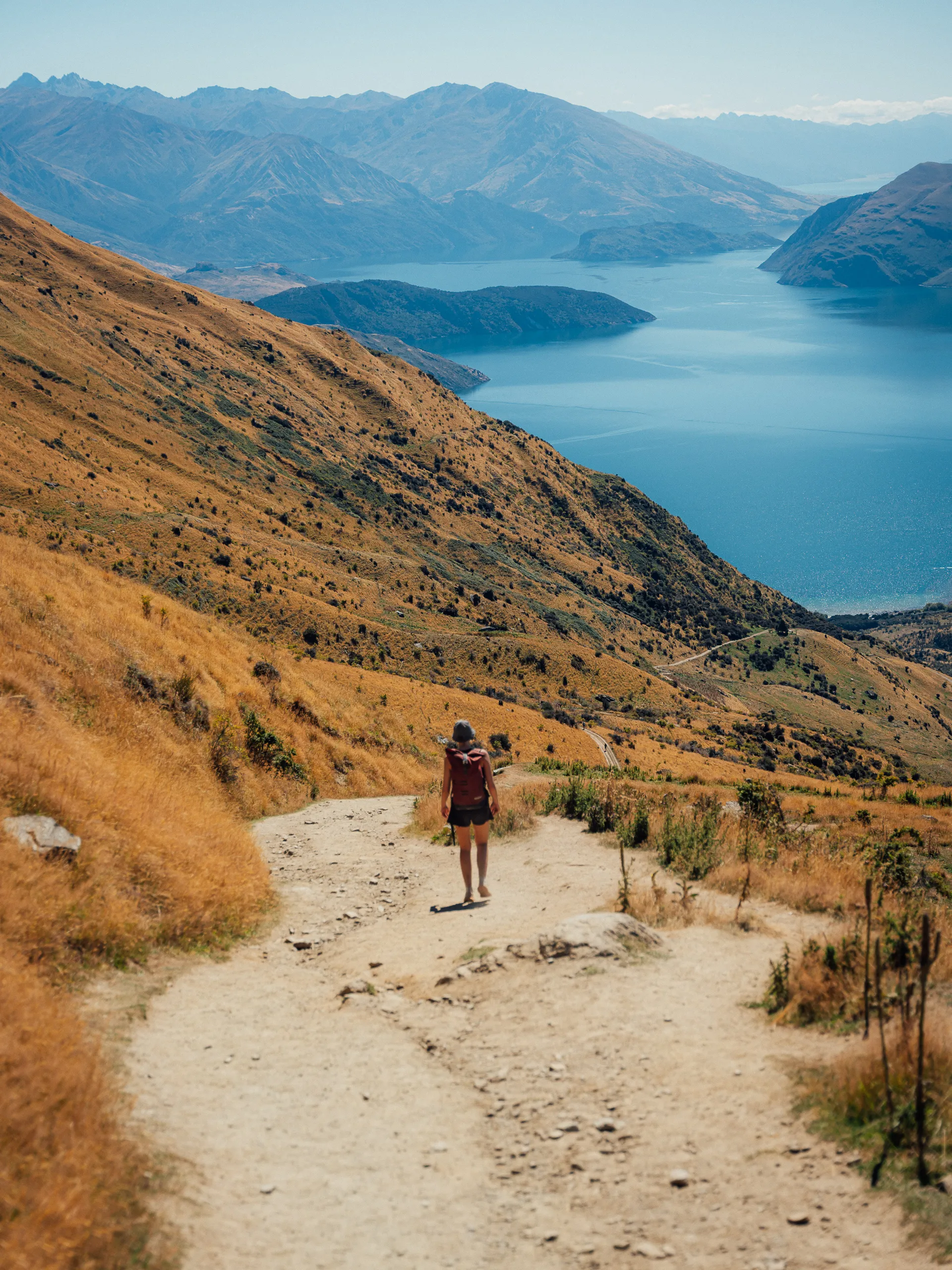 A person with a backpack walking down a steep, winding dirt path on a golden-brown hillside with mountains and Lake Wānaka in the background.