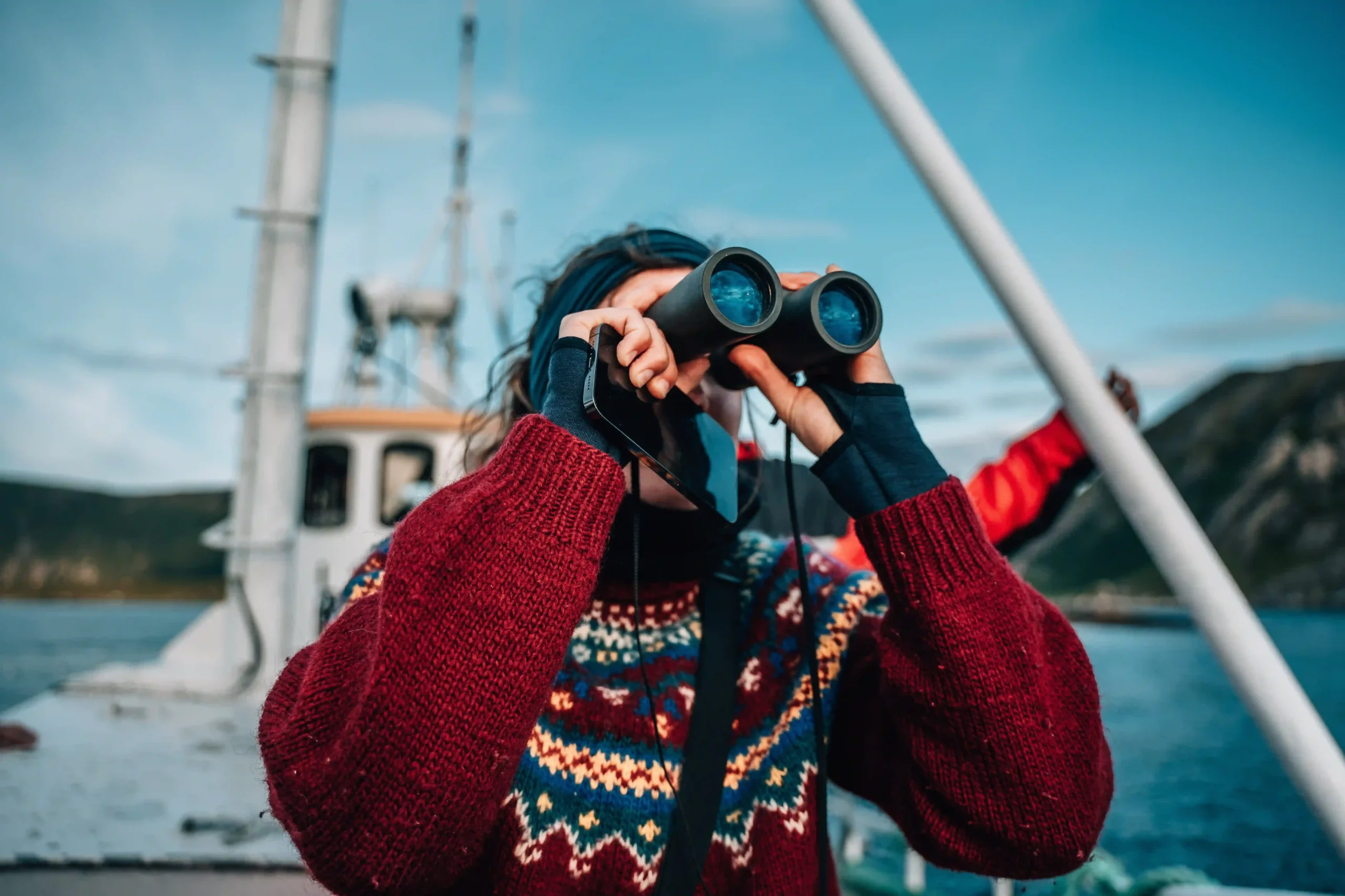 A person in a red patterned Nordic sweater looking through black binoculars from the deck of a boat.