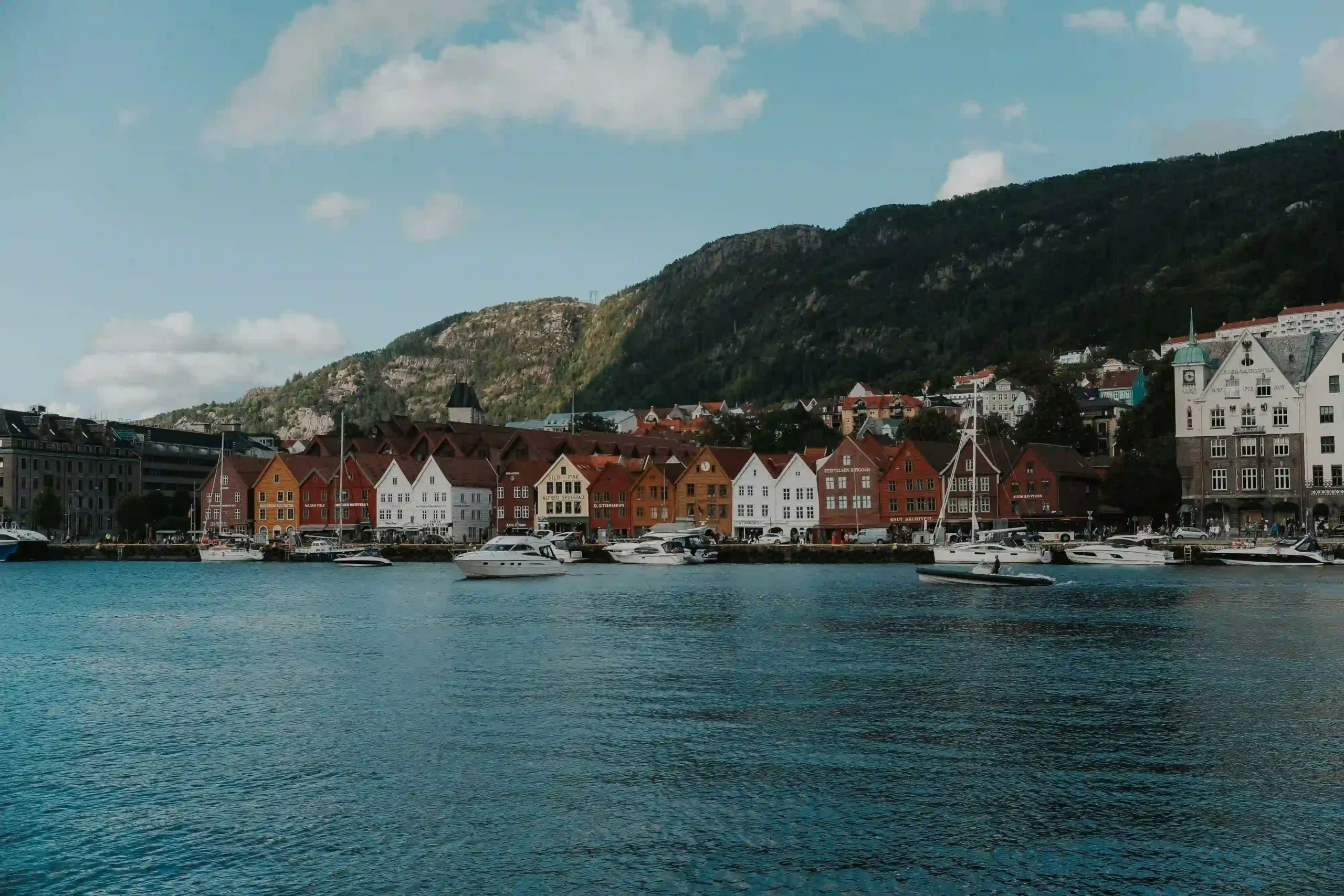 A wide view of the famous Bryggen wharf in Bergen, Norway. A row of historic, colorful wooden houses in red, white, and yellow stands along the waterfront. Several boats are docked in the blue water in front of the buildings, with a steep, green mountain rising in the background under a blue sky.