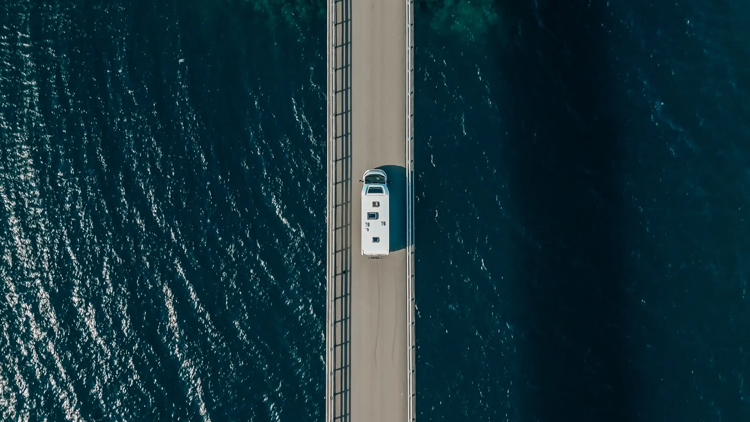 A vertical top-down aerial shot of a white motorhome driving along a narrow bridge that cuts through deep, dark blue seawater in Norway.