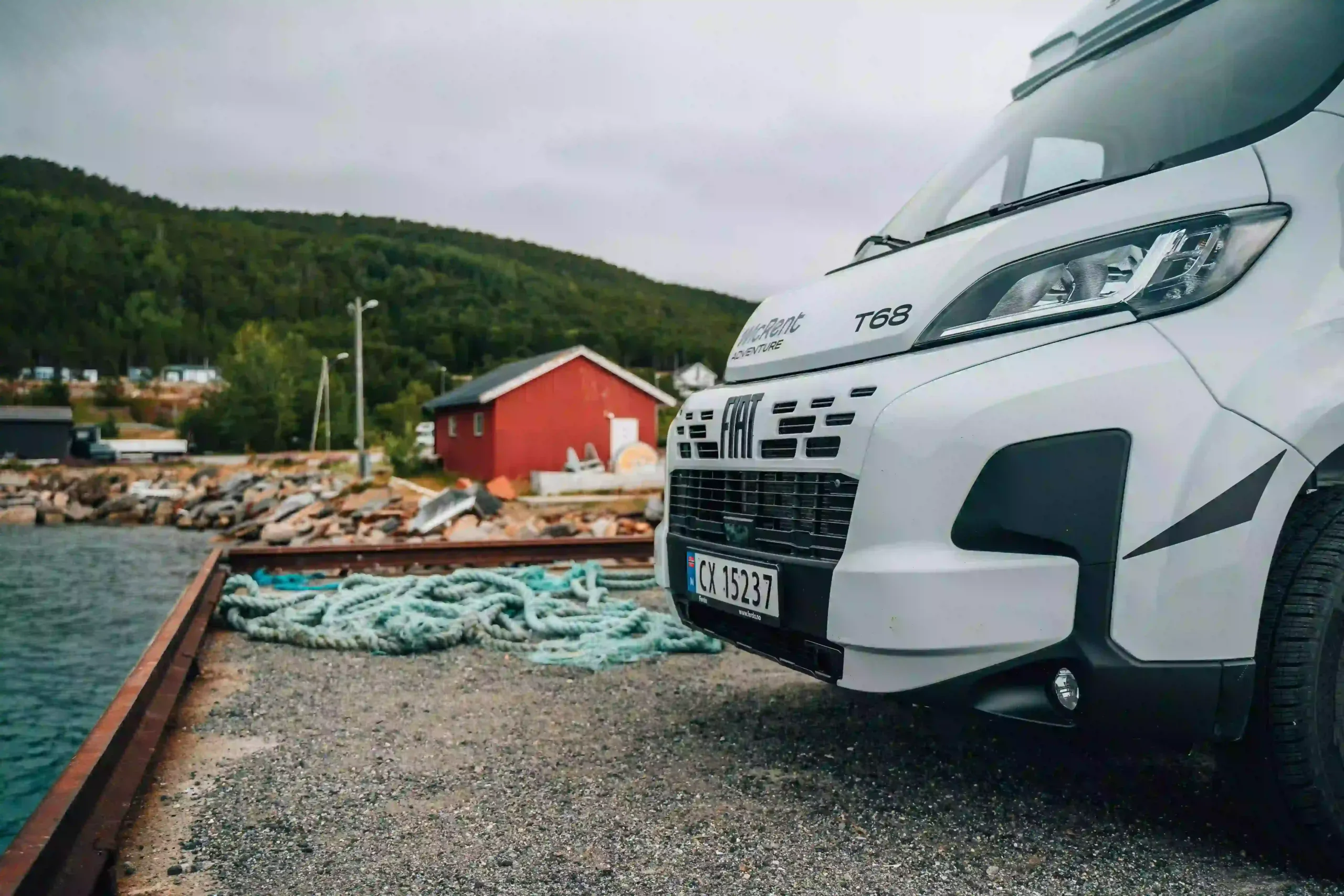 A close-up, angled view of the front of a white campervan parked on a gravel pier. In the background, there is a traditional red wooden Norwegian hut, a pile of turquoise fishing nets, and lush green hills.