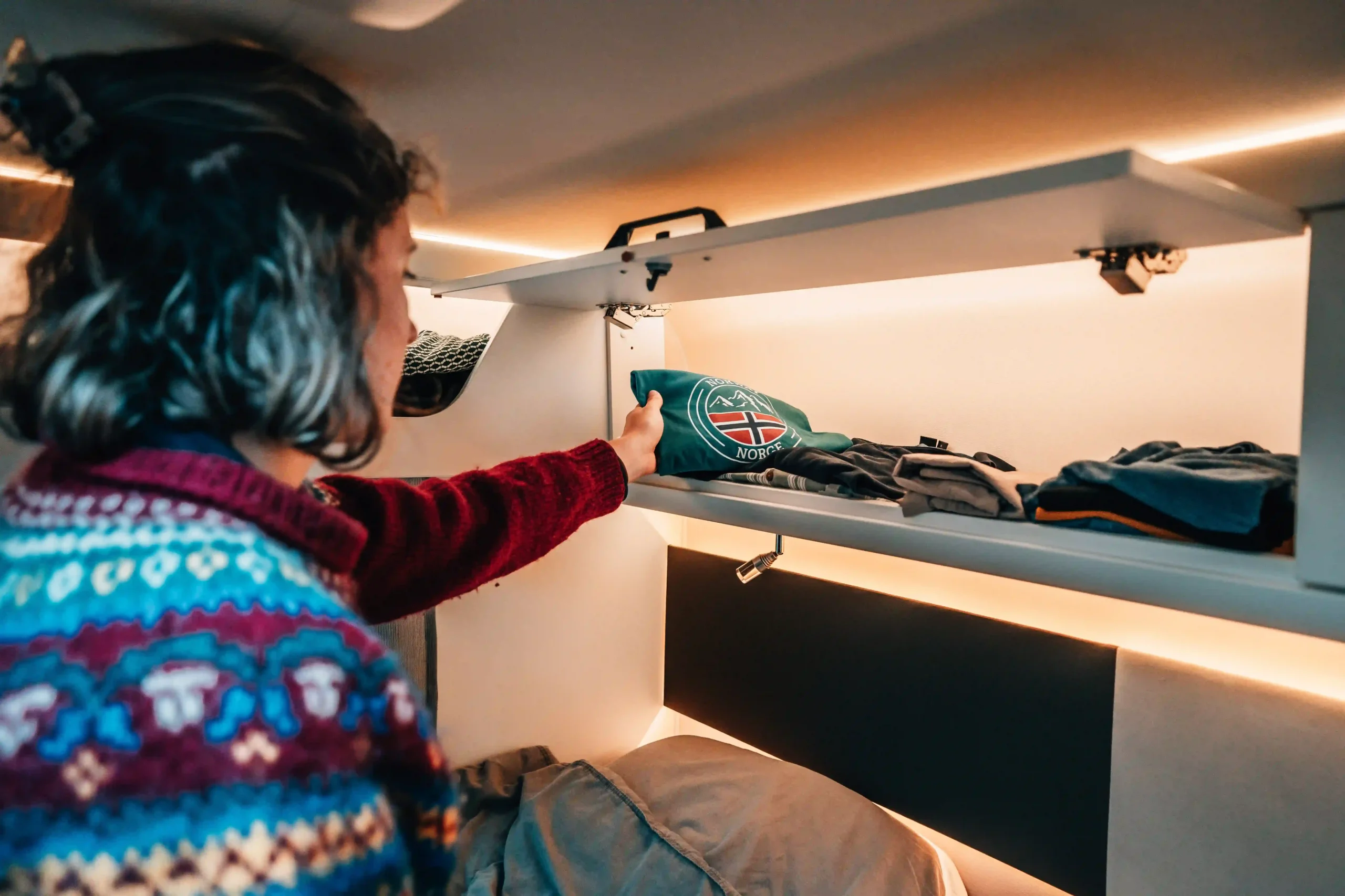 An interior view of a campervan. A person wearing a colorful patterned wool sweater reaches into an overhead storage cabinet.