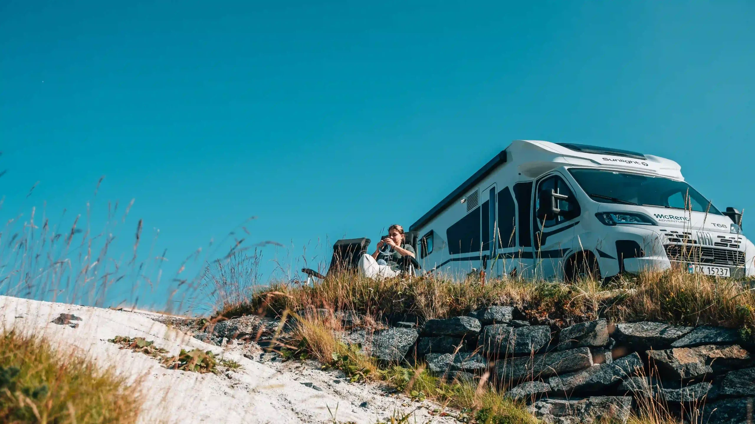 A low-angle shot looking up a grassy, rocky slope at a white campervan parked on a ridge.