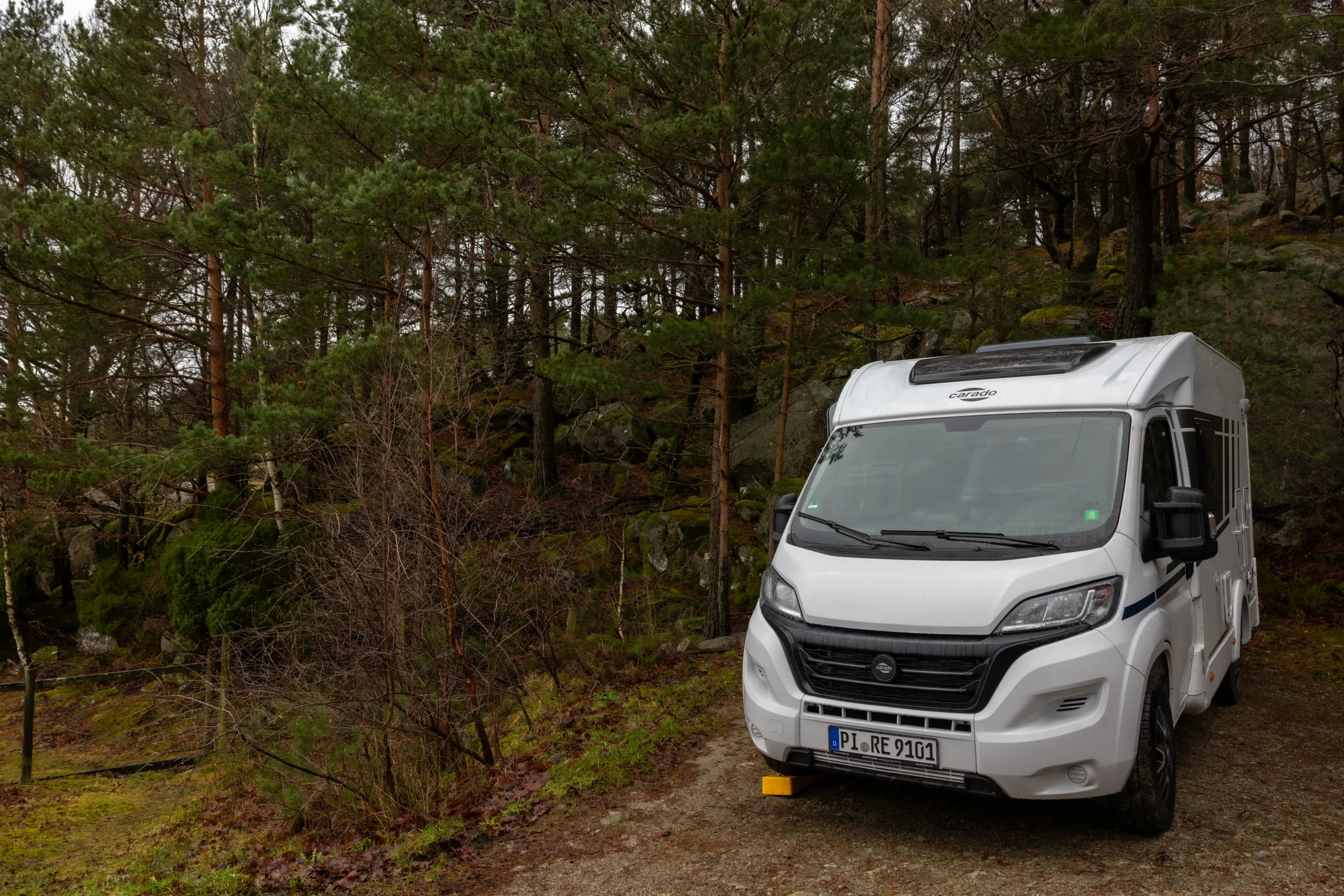 A white motorhome parked on a gravel path at the edge of a dense Norwegian forest with moss-covered rocks.