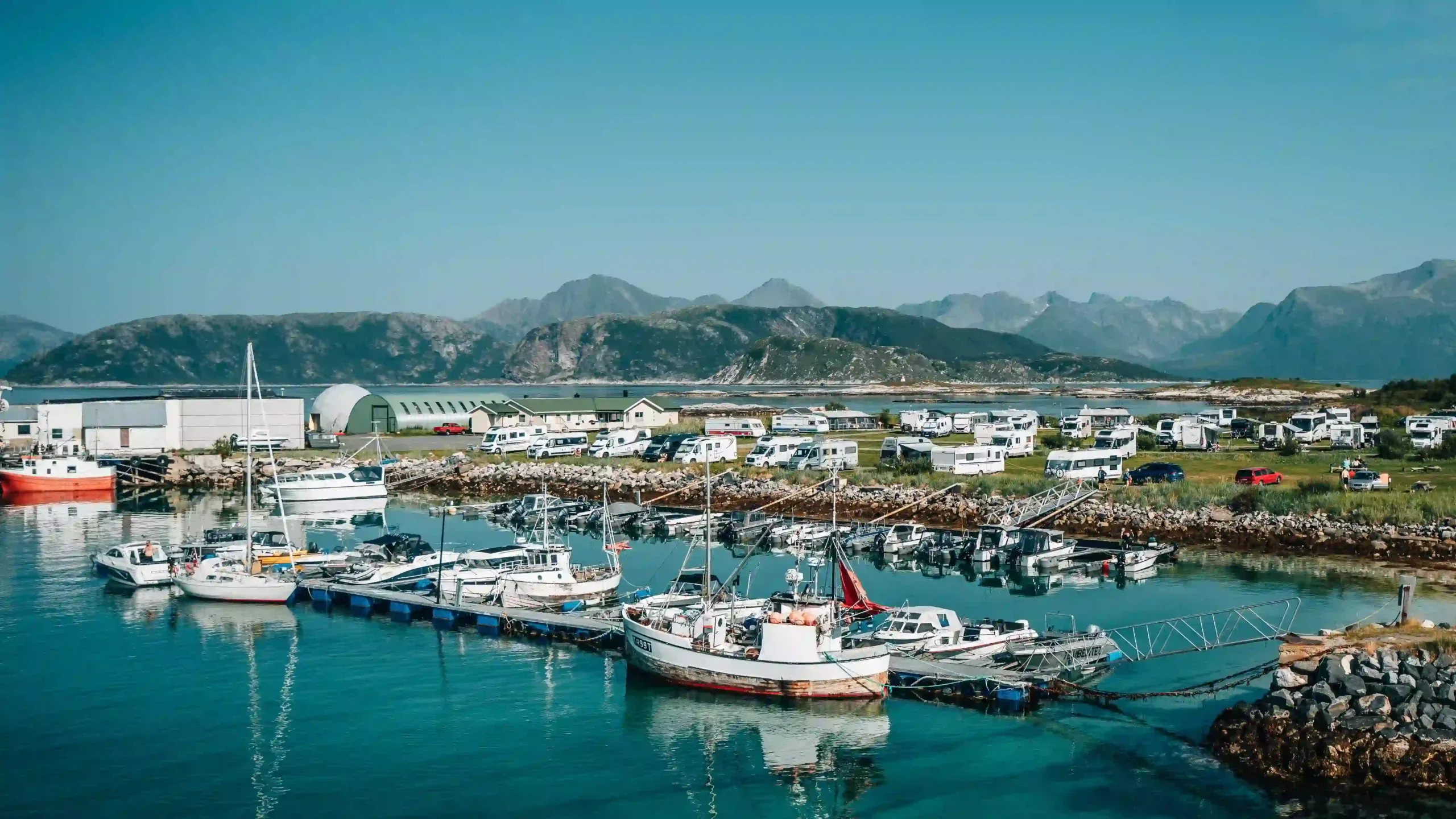 High-angle landscape view of boats docked in a clear turquoise harbour, with a busy motorhome campsite along the green shoreline and rugged mountains in the background.
