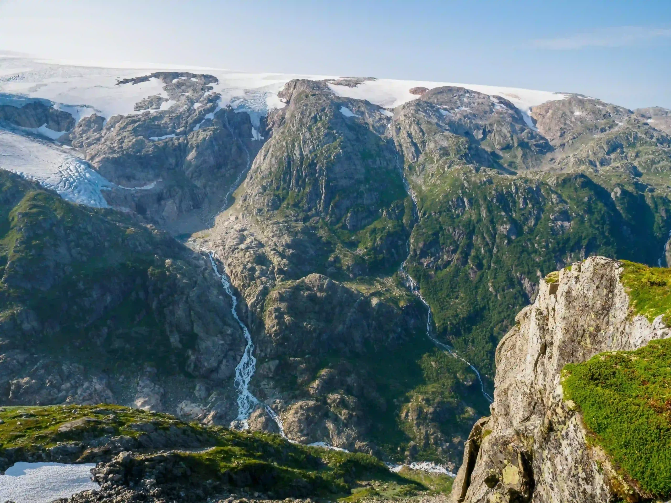 High-angle view of a massive white glacier ice cap resting on rugged, rocky mountain peaks with cascading meltwater waterfalls.