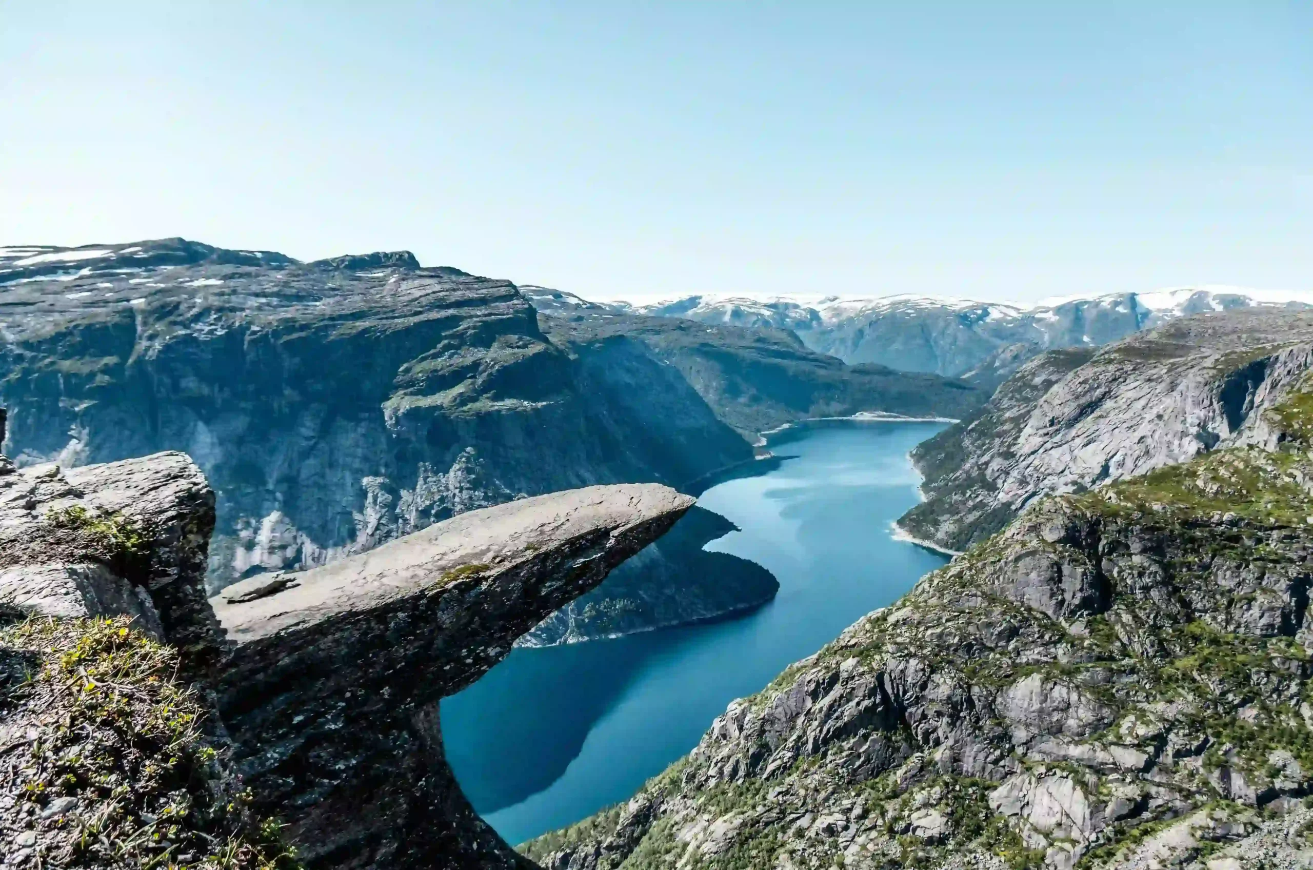 A dramatic rock plateau known as Trolltunga sticks out horizontally from a mountain cliff in Norway. Below the rock is deep blue water, surrounded by steep, rugged mountains under a clear blue sky.