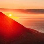 A wide landscape photograph of a rugged mountain slope meeting the sea during a vibrant sunset.