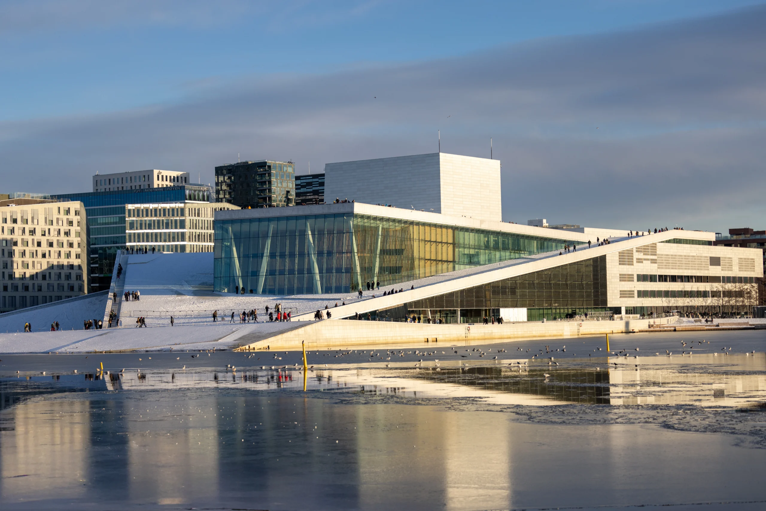 The white, angular marble architecture of the Oslo Opera House covered in snow, with people walking on the sloping roof.