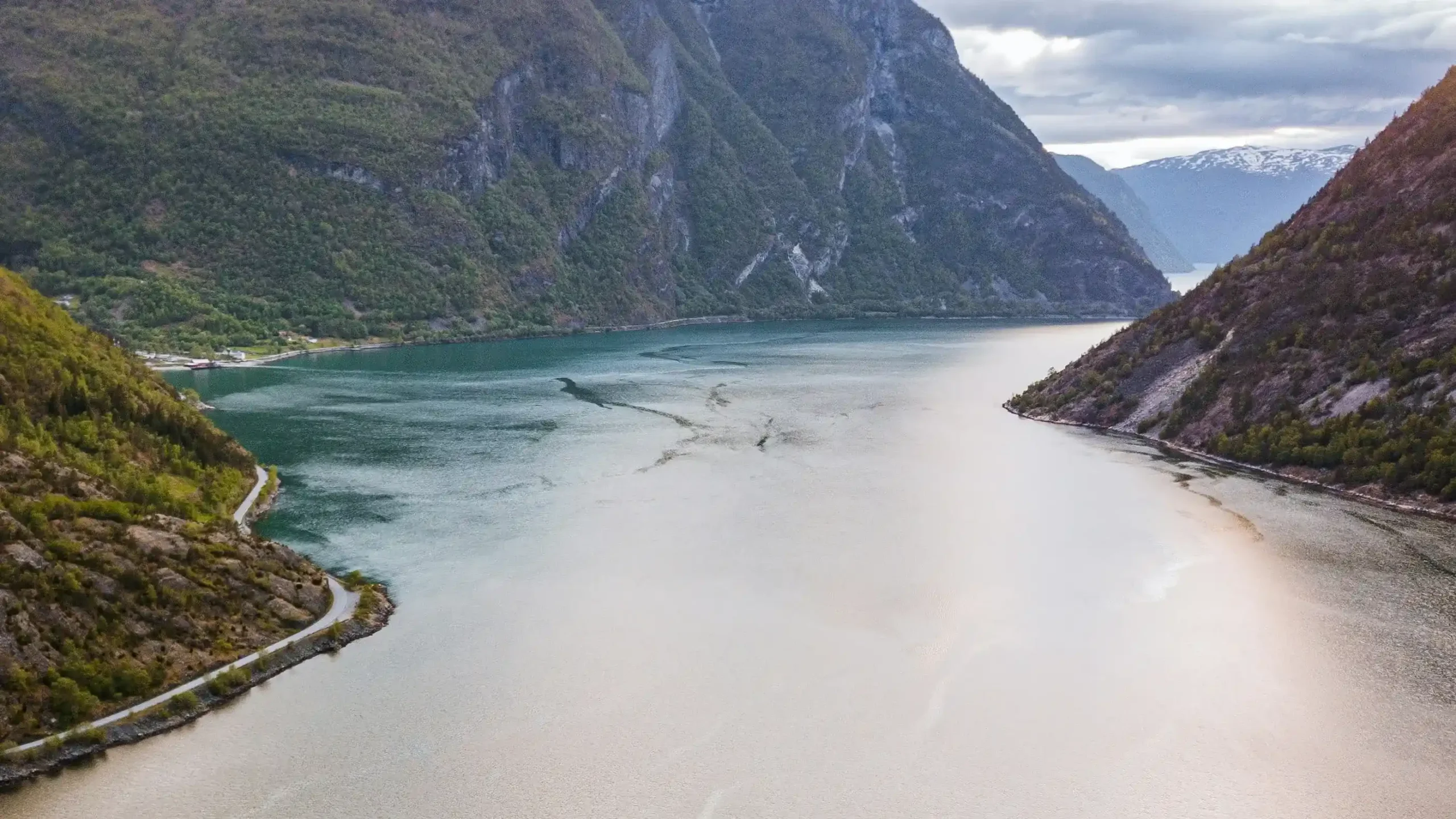 High aerial view of Sognefjord in Norway. Calm water, framed by steep green mountains and a winding fjord-side road.