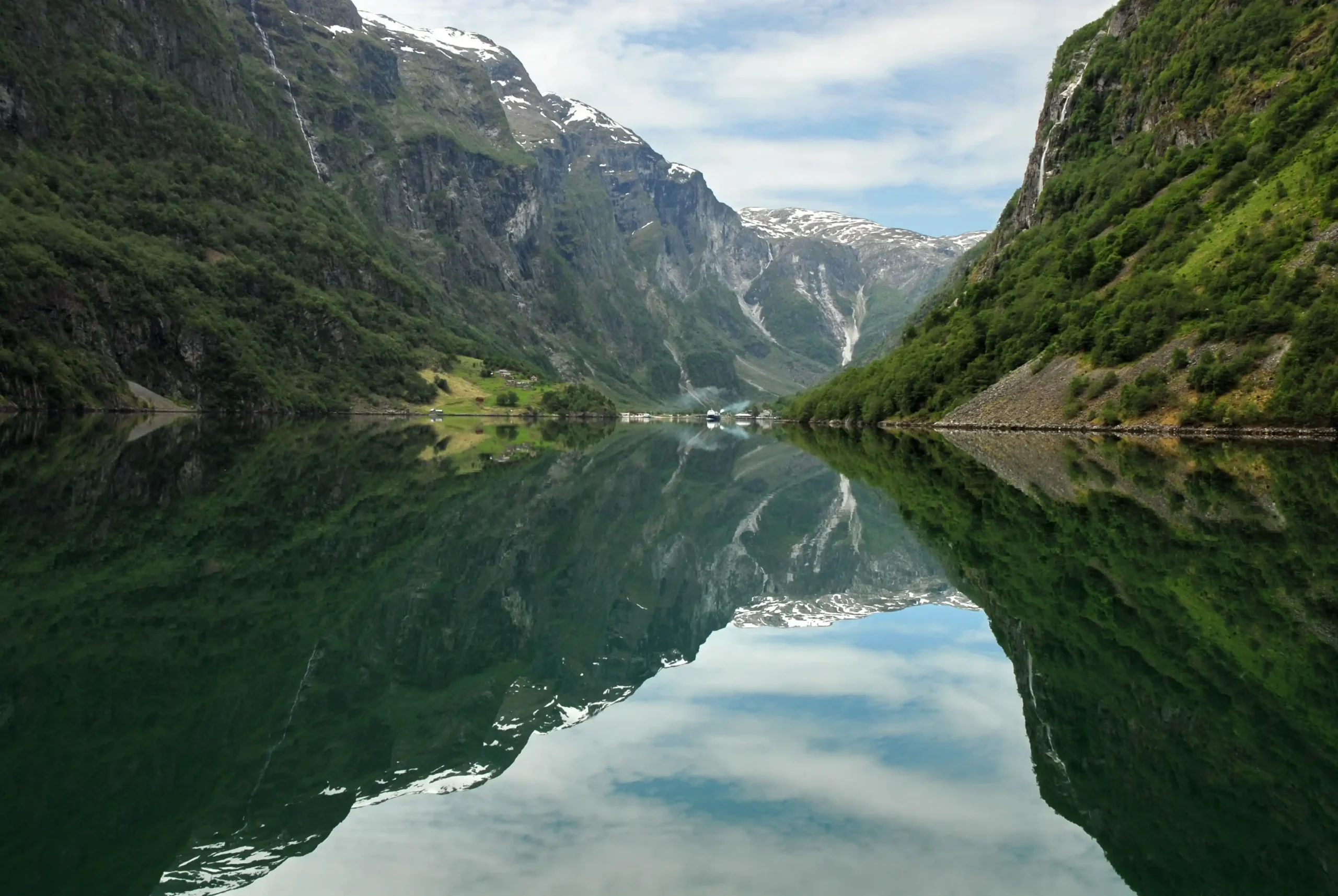 Reflection of steep, green glacial mountains and snow-tipped peaks in the calm, emerald-green water of a narrow fjord.