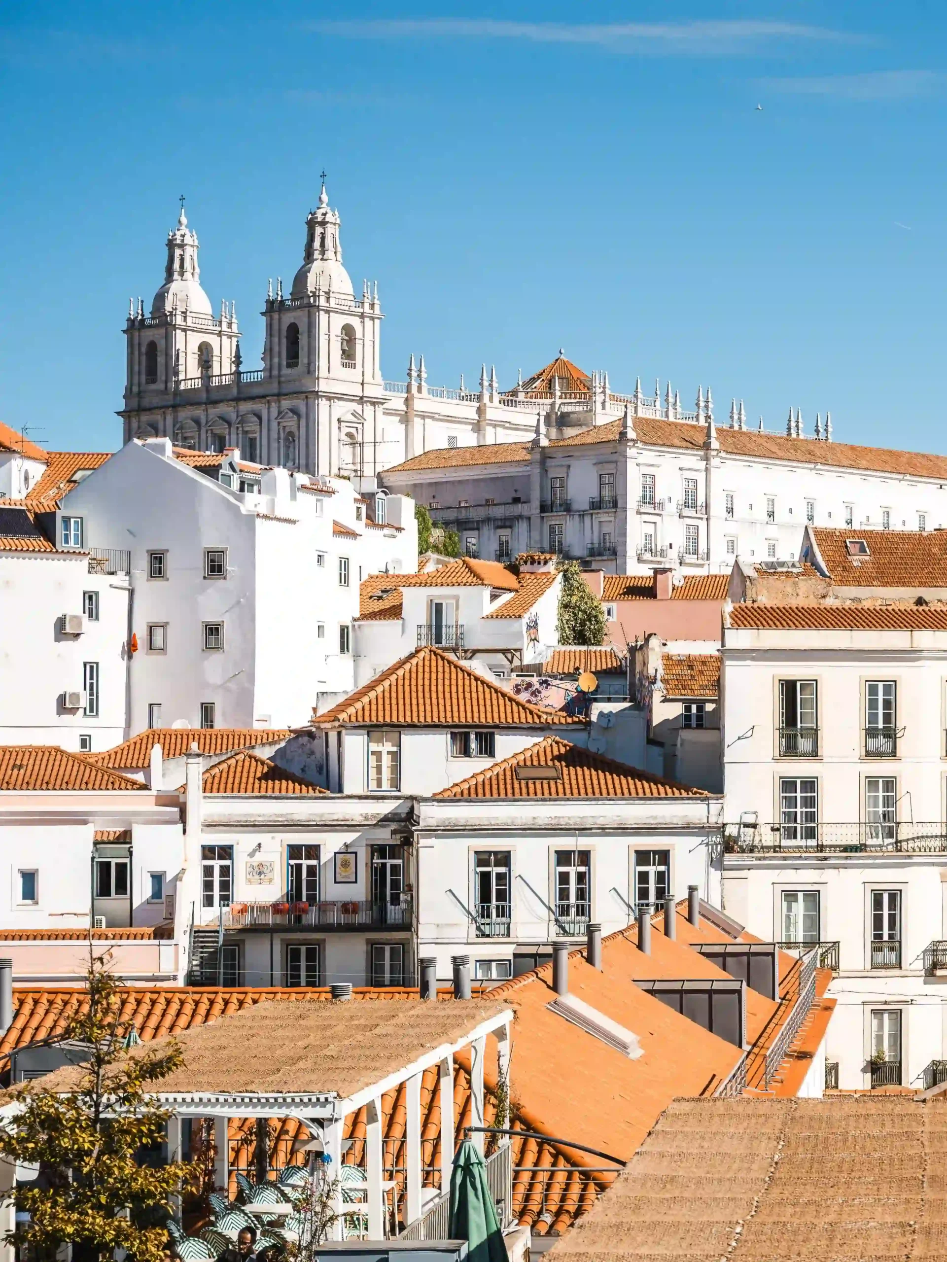 View looking over the dense white buildings and orange terracotta tiled roofs of Lisbon, Portugal. In the background, the grand white towers of the Church of São Vicente de Fora stand out against a clear, vibrant blue sky.